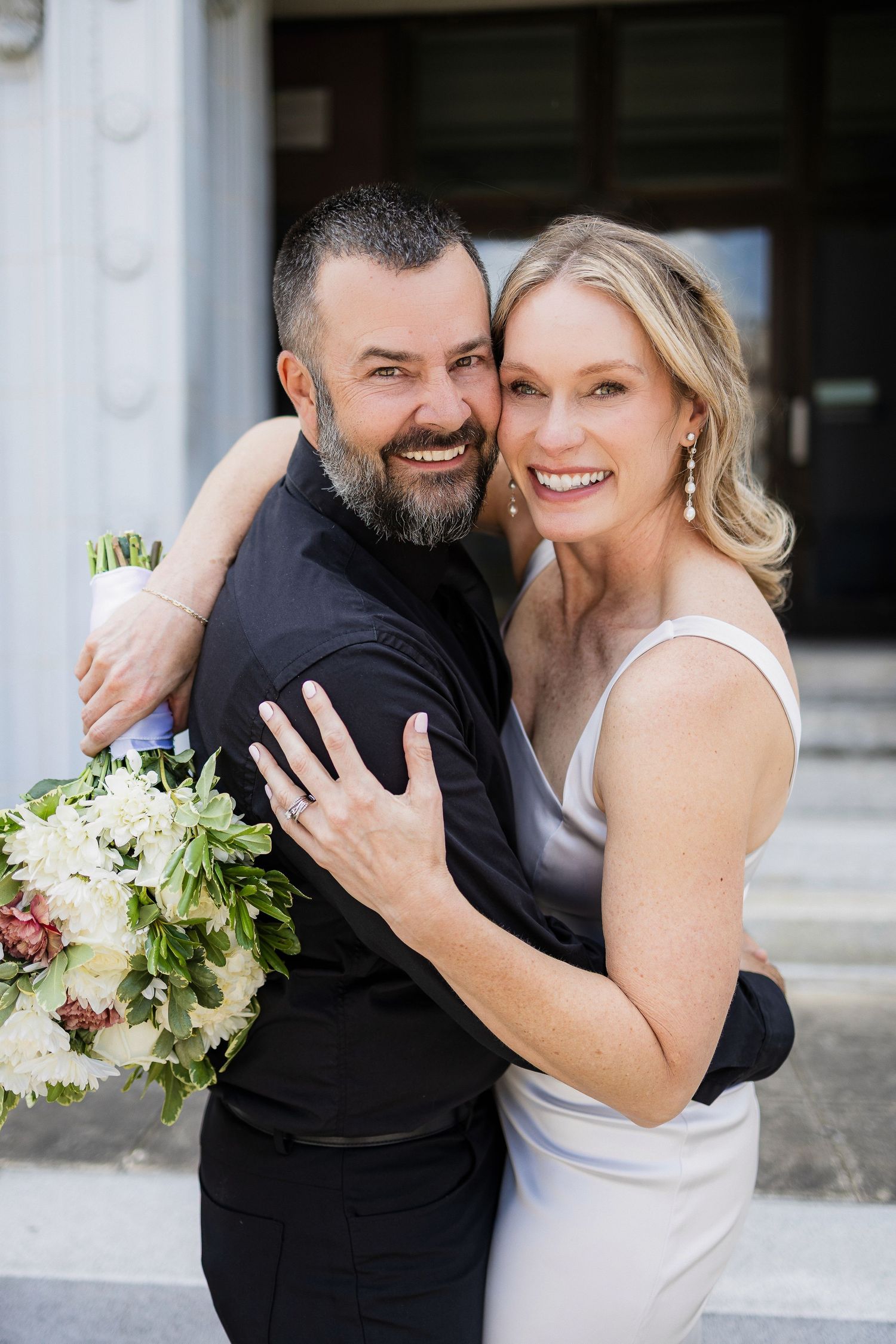 A joyful wedding couple laughs and embraces while holding a white floral bouquet outdoors.