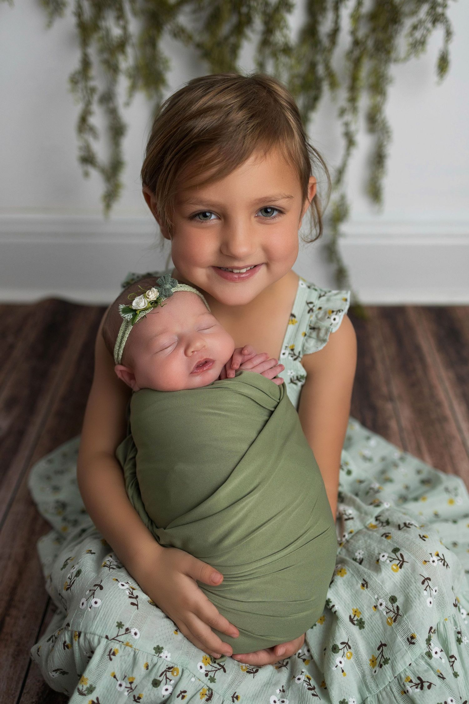 Baby being held in green wrap by someone wearing floral dress against light studio backdrop.