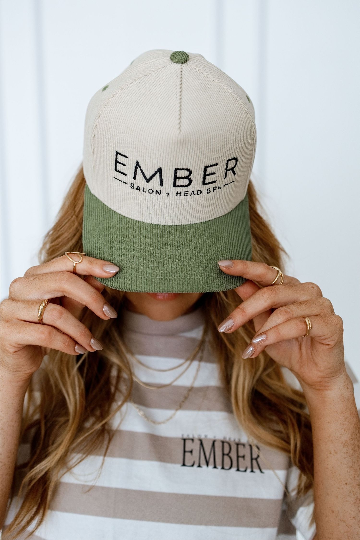 Woman holding an Ember Salon and Head Spa two-tone green and beige snapback hat over her face.