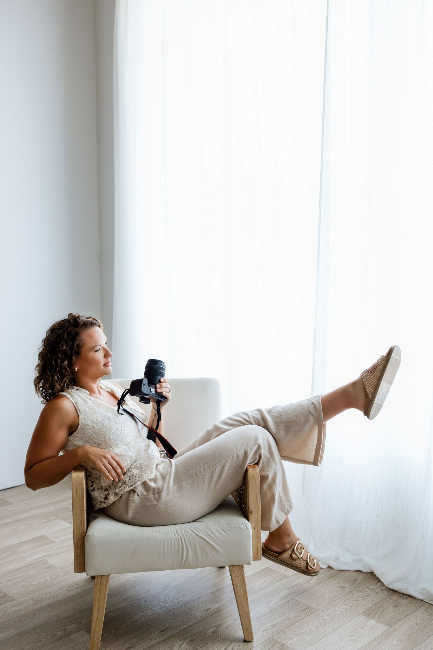 Woman with curly hair relaxing in armchair holding DSLR camera near bright white curtained window.
