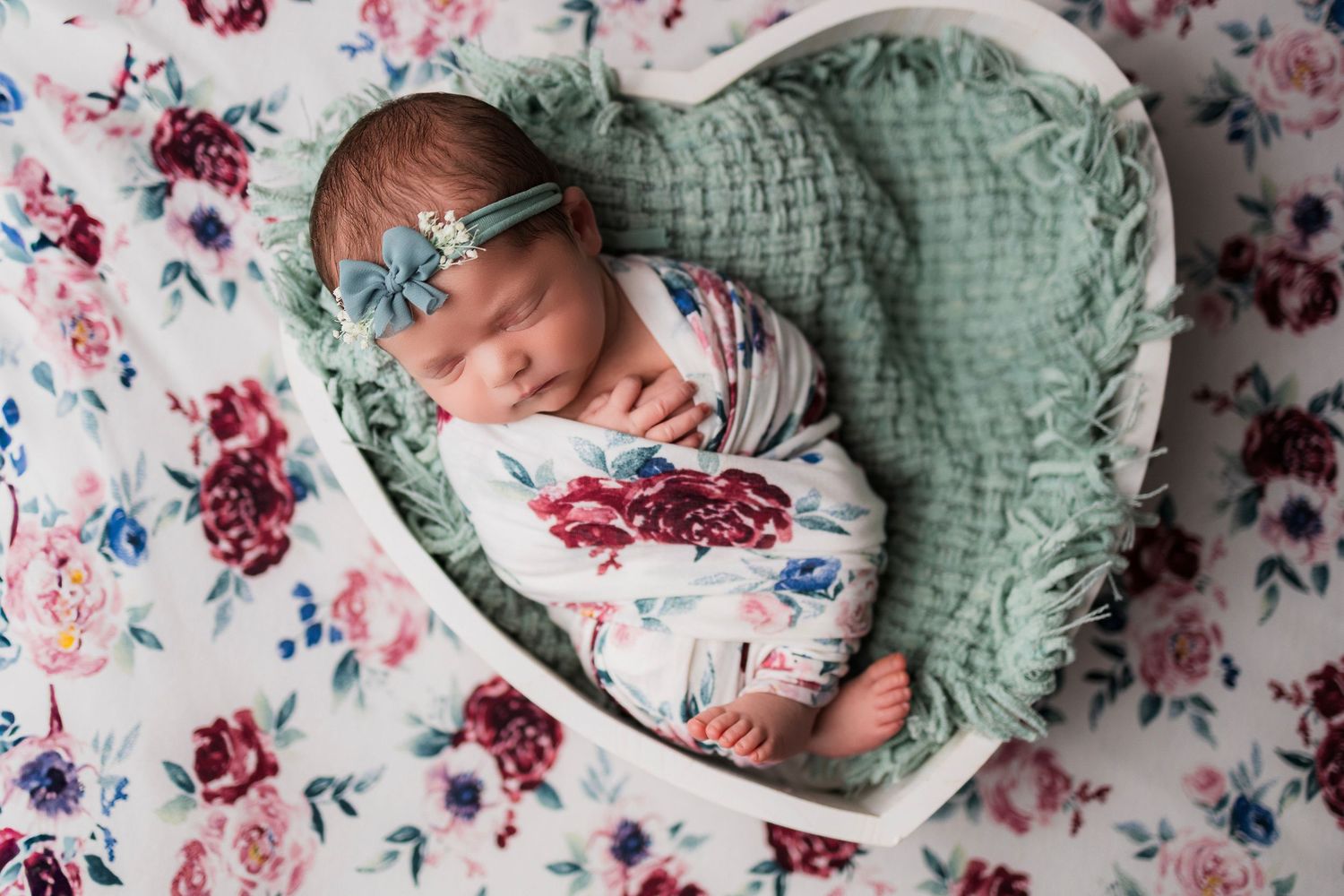 Newborn baby sleeping peacefully in heart-shaped basket on floral blanket with sage green knit blanket.