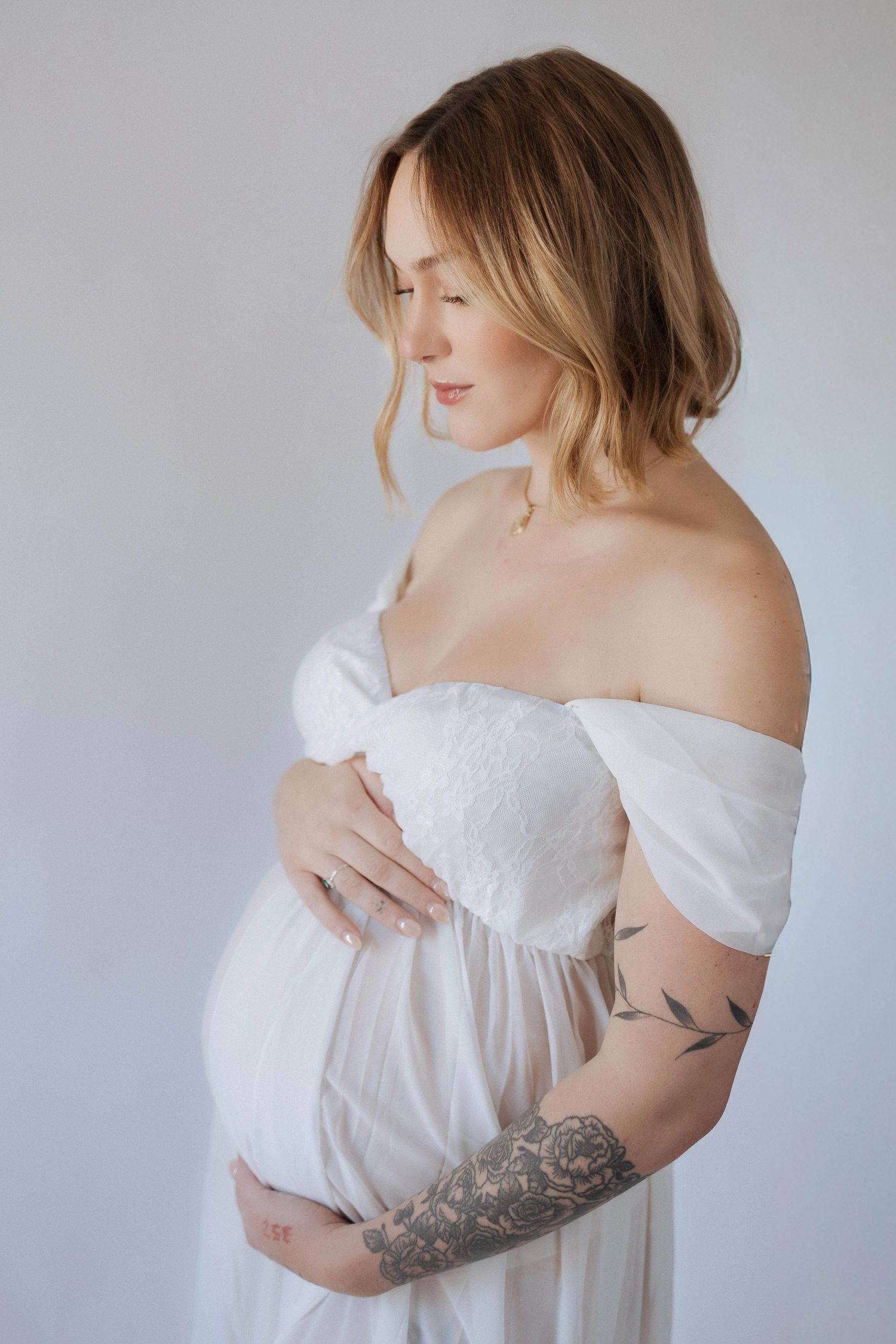 Maternity portrait in white flowing off-shoulder dress showing tattoo on arm against light gray backdrop.