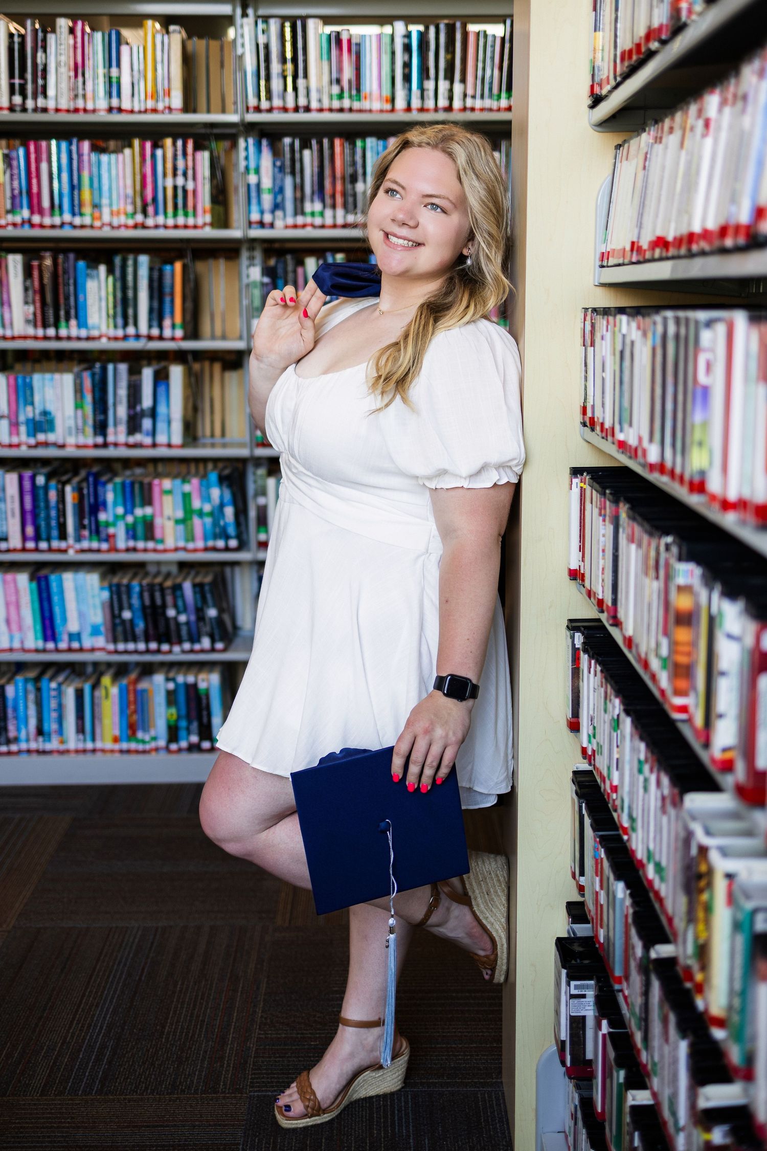 Graduate in white dress poses cheerfully between library bookshelves holding diploma.