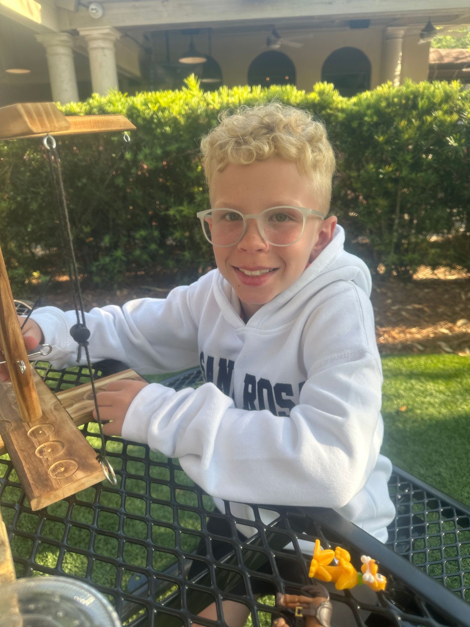 Young person in white hoodie smiling at outdoor patio table with greenery in background.
