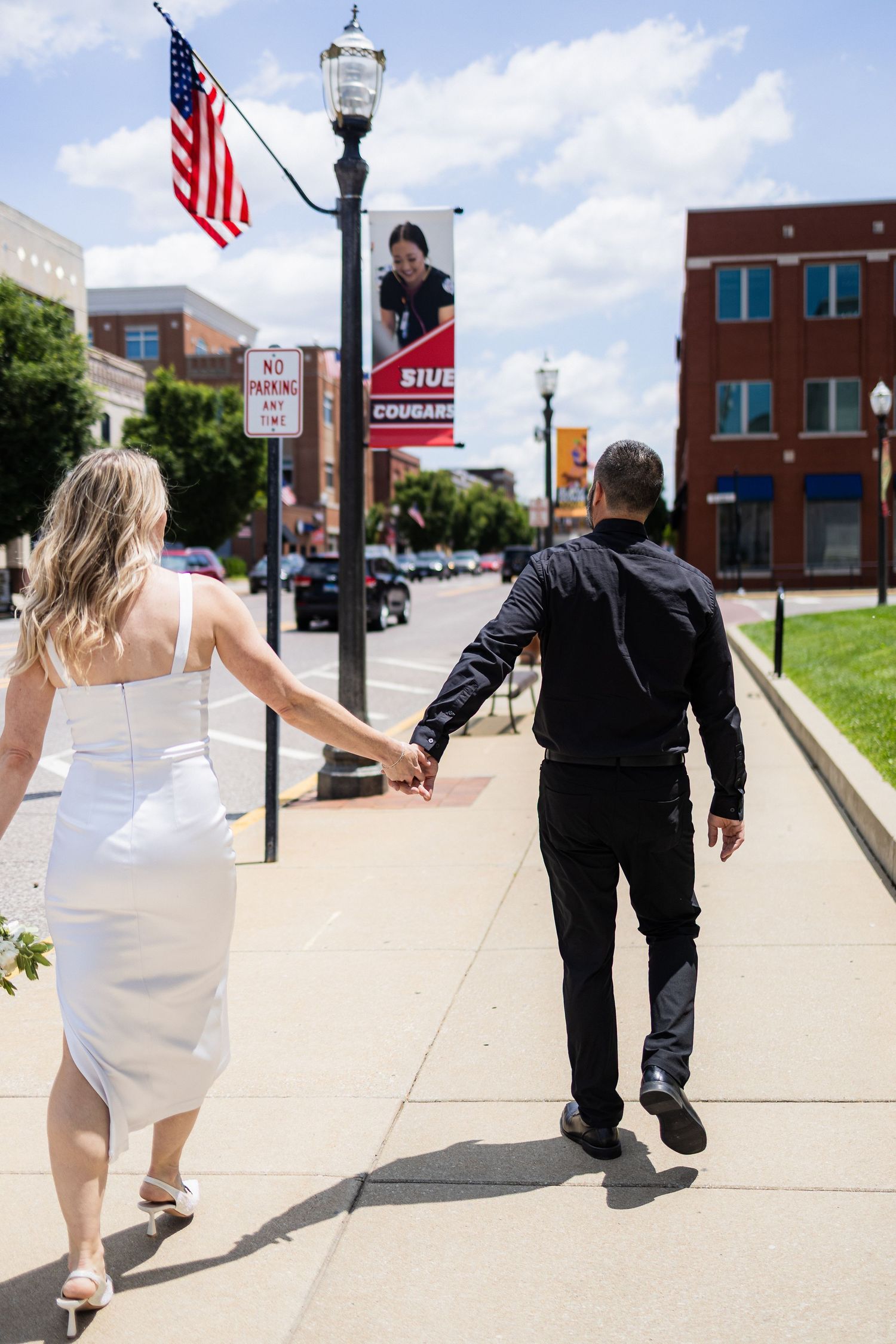 Couple walking on a city sidewalk holding hands, with an American flag and lamp post visible in the background.