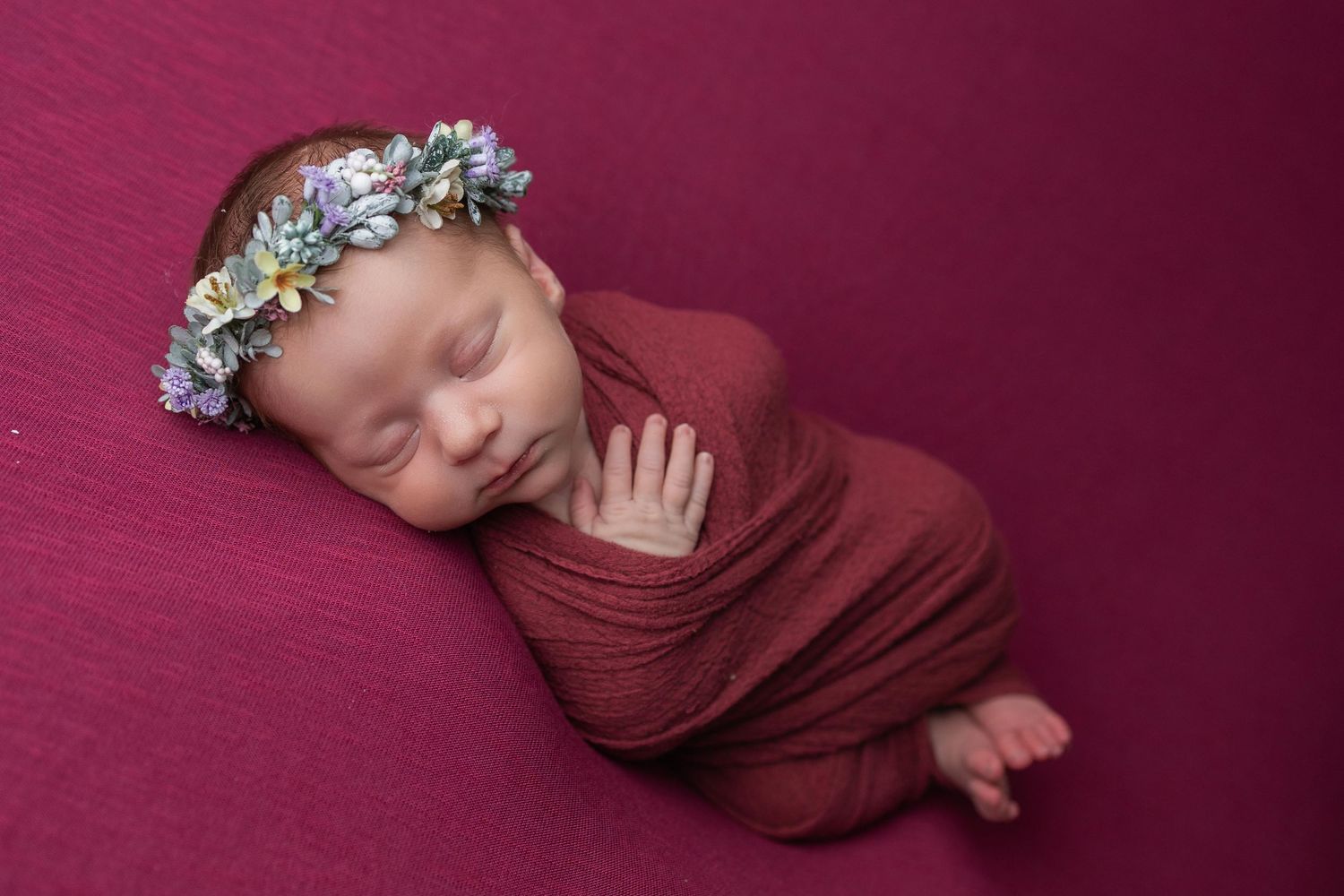 A newborn baby sleeps peacefully wrapped in a burgundy swaddle with delicate floral hair accessories against a pink backdrop.