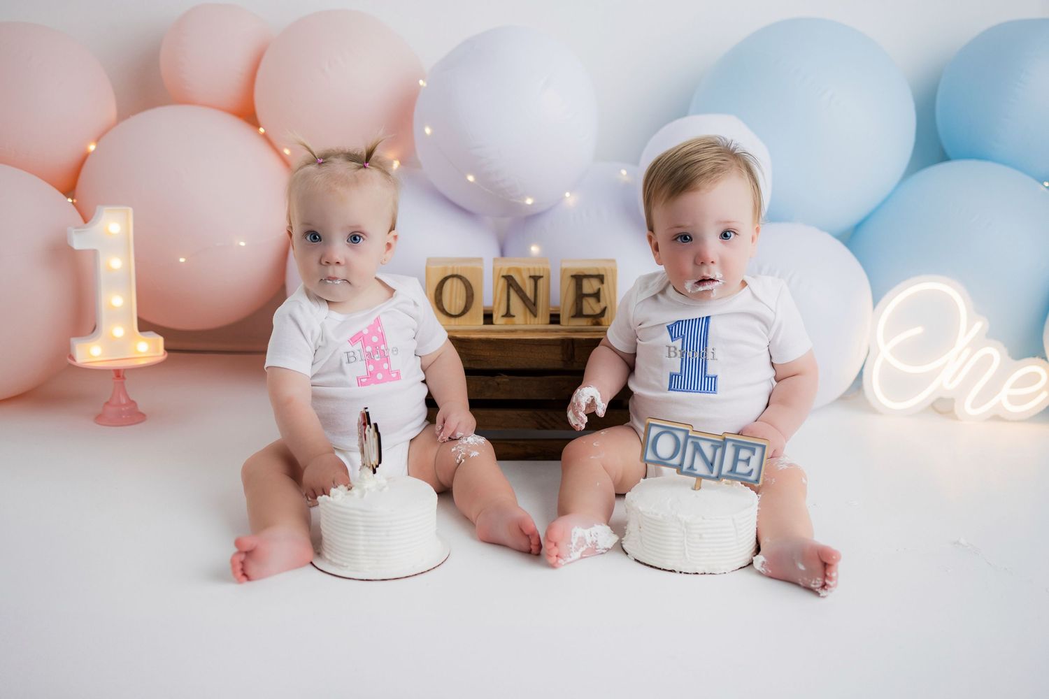 Two babies sit beside lit number one candles and balloons during a first birthday celebration photo shoot.