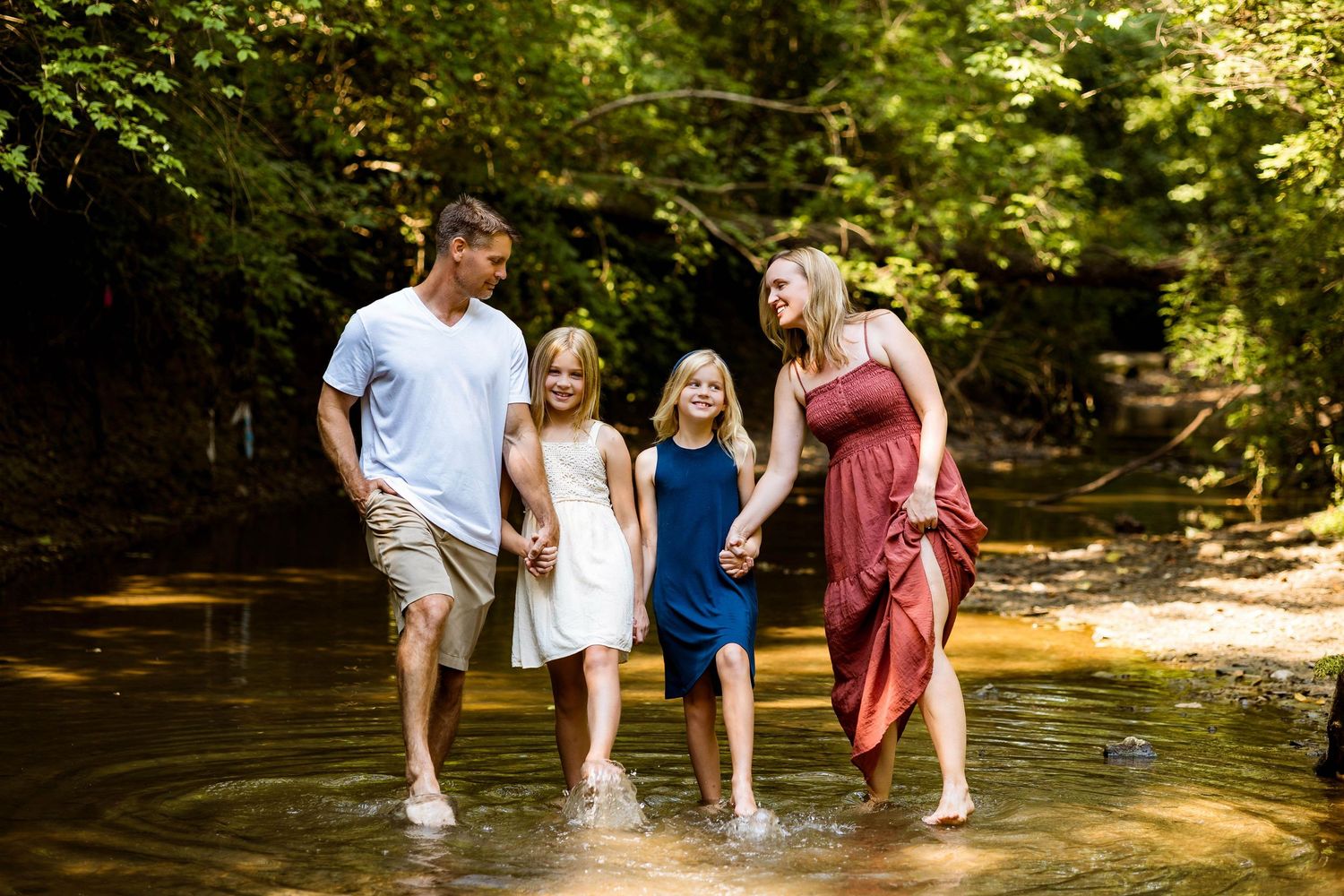 A family splashes together in shallow creek water on a warm summer day amid sunlit trees and forest scenery.