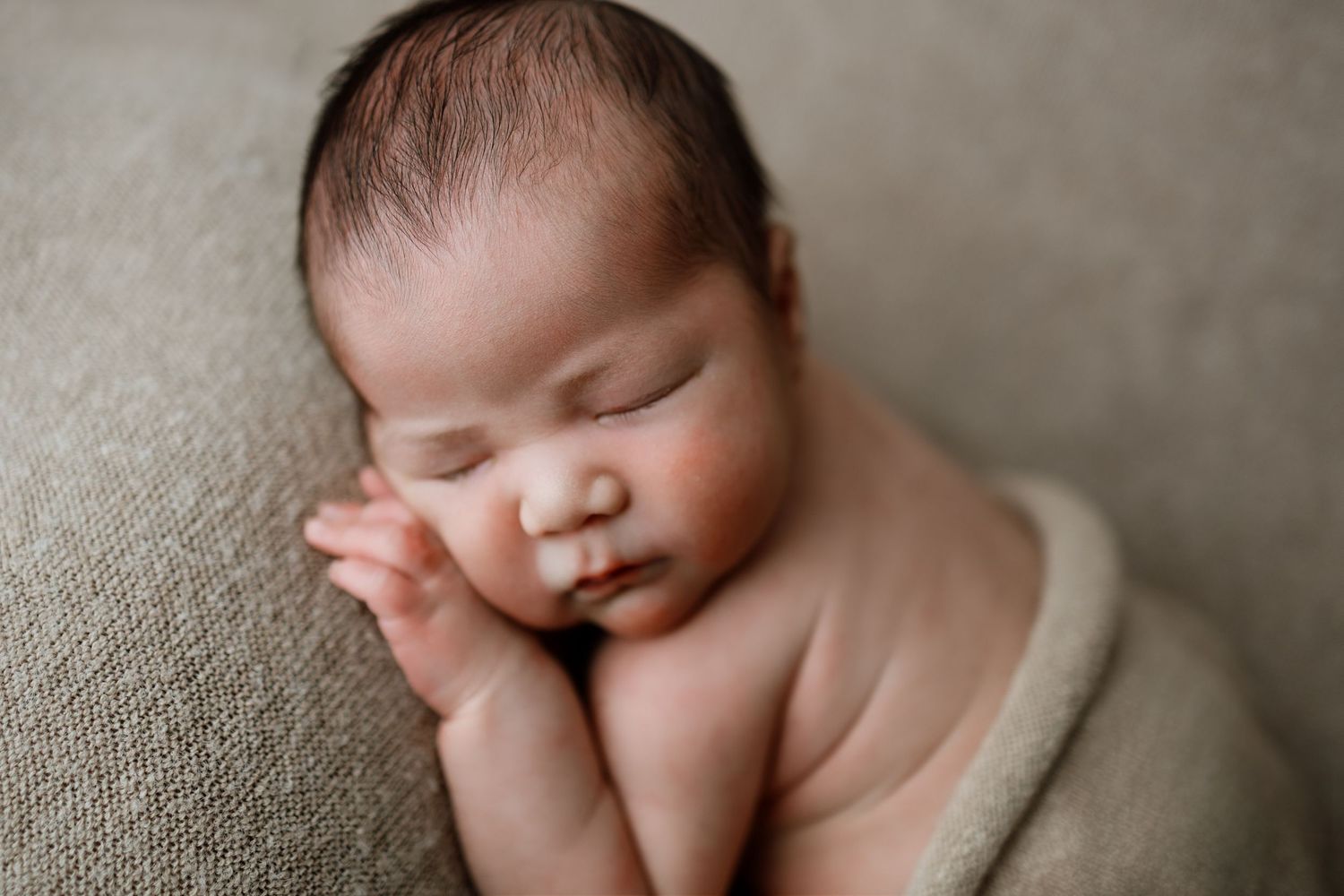 Sleeping newborn baby with peaceful expression rests on textured gray blanket in soft lighting.