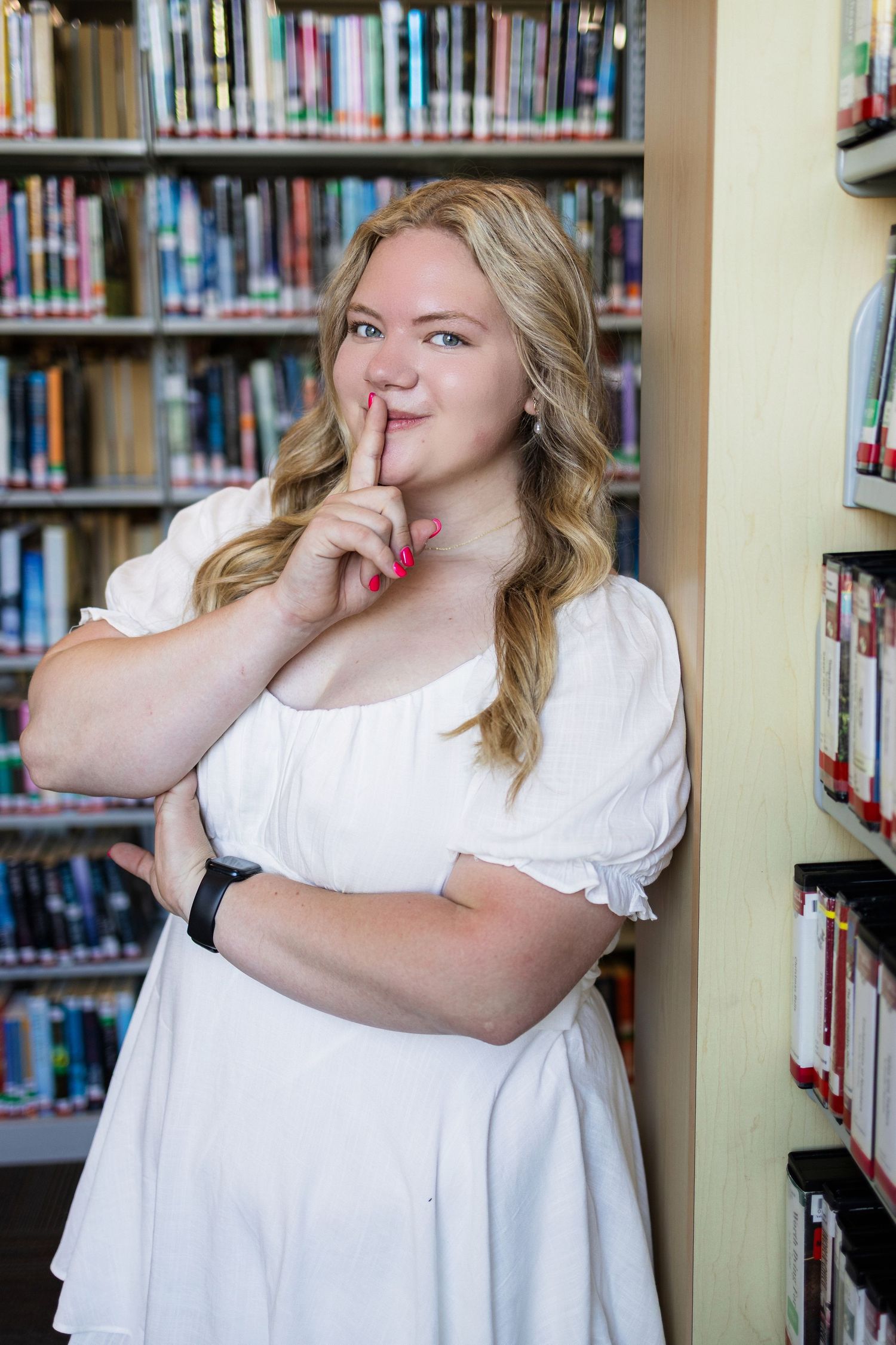 Graduate in white dress makes playful shushing gesture while posing in library with bookshelves in background.