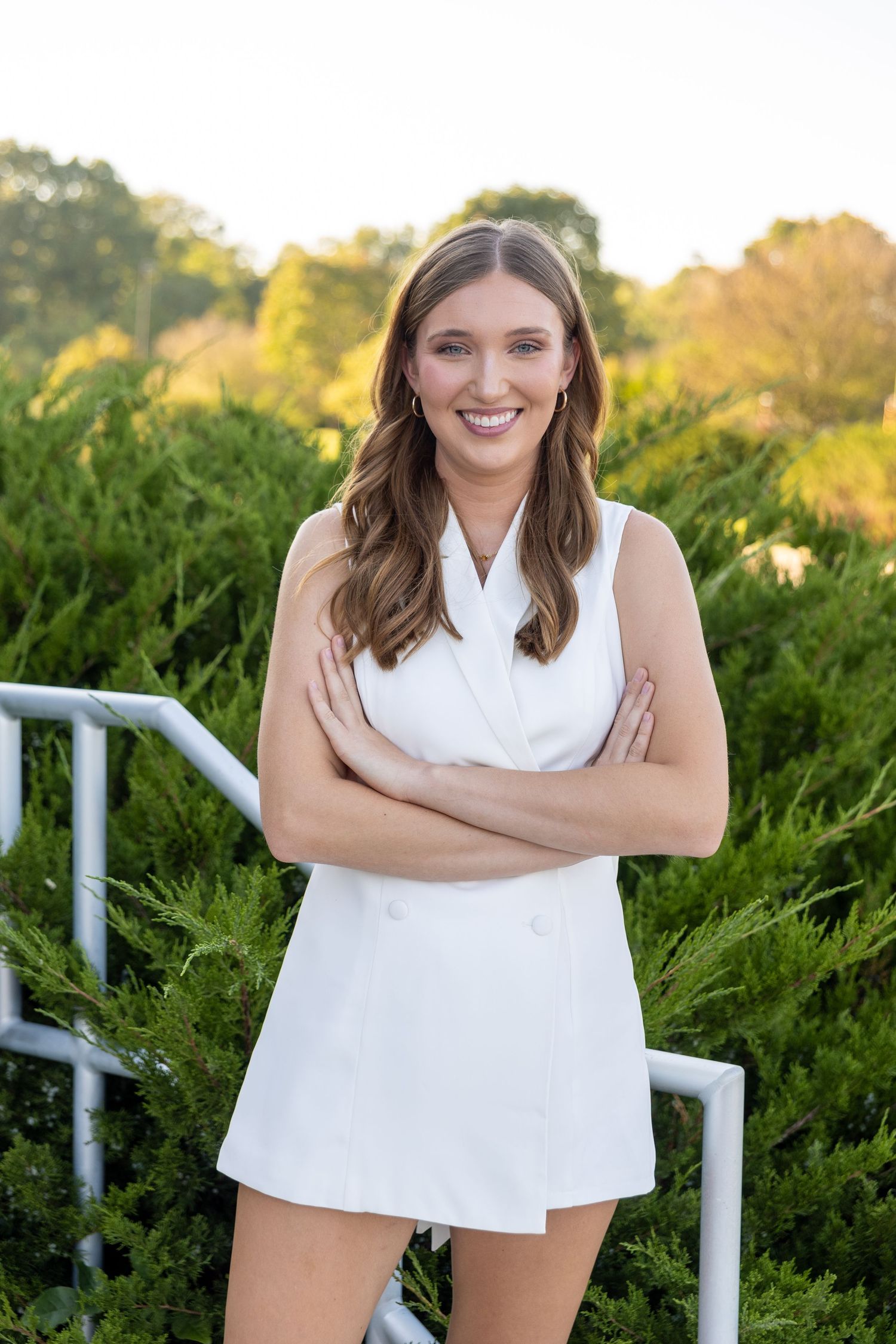Person in white dress poses confidently with crossed arms against lush outdoor garden setting at sunset.