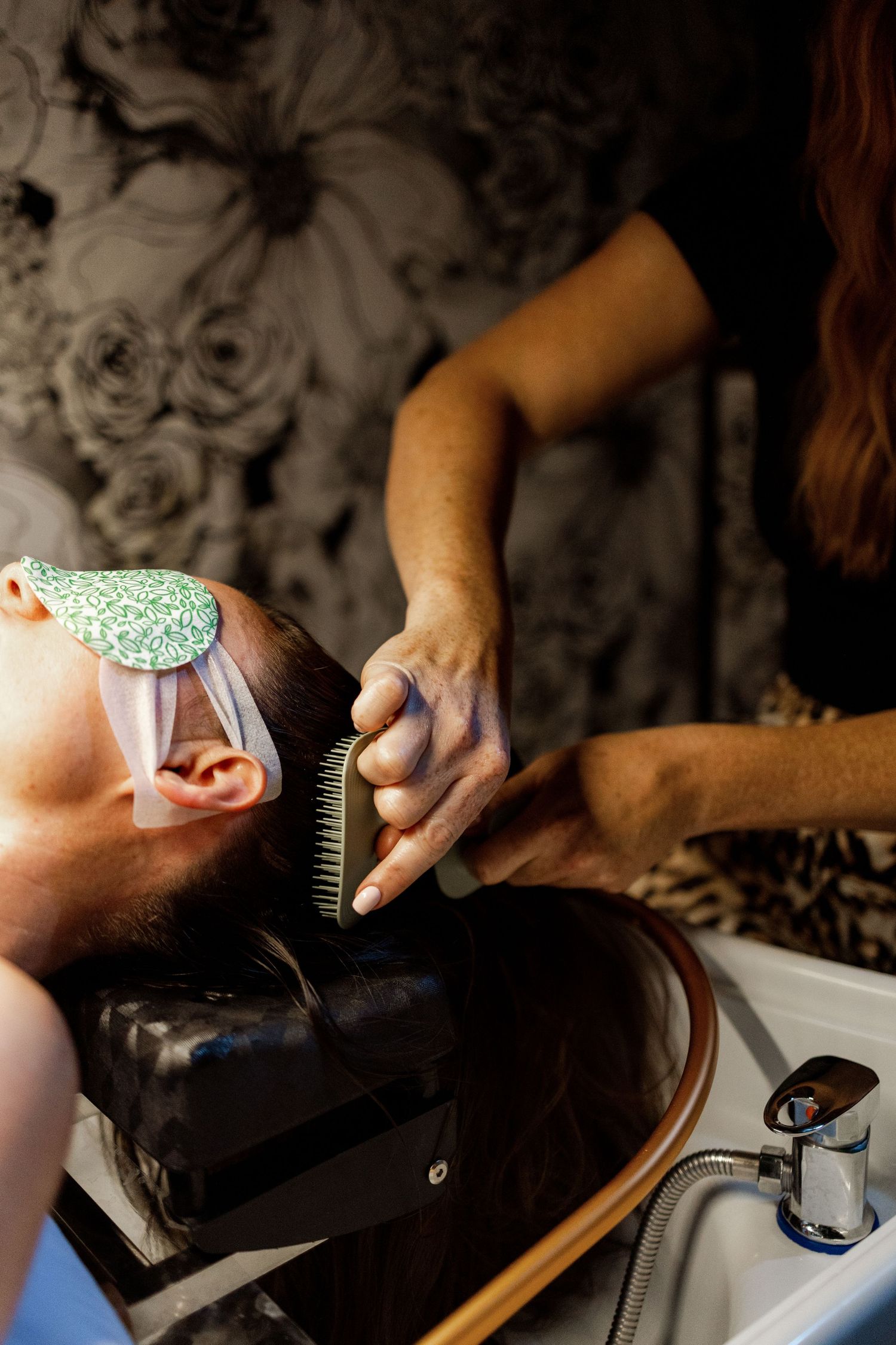 Hair stylist brushes a client's hair at a salon wash basin during bridal wedding day preparations.