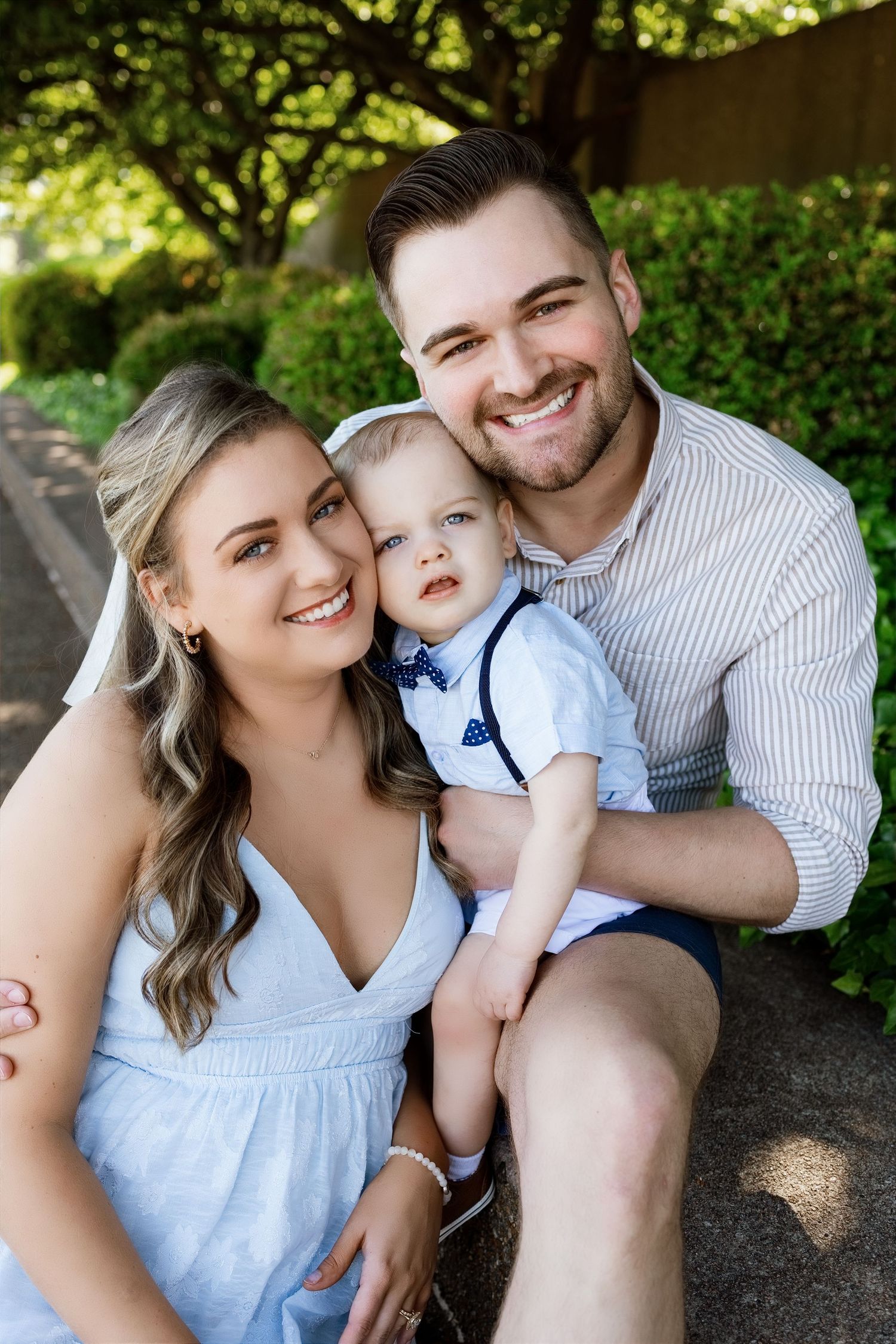 Young family smiling outdoors, parents holding toddler boy in bow tie, seated in lush green garden setting.