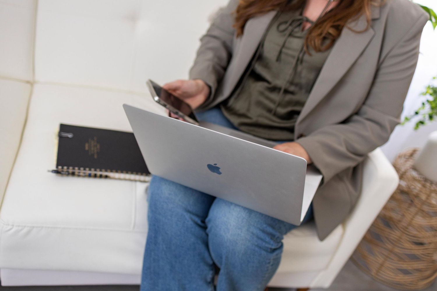 Person in grey blazer working with MacBook laptop and smartphone while sitting on white chair.