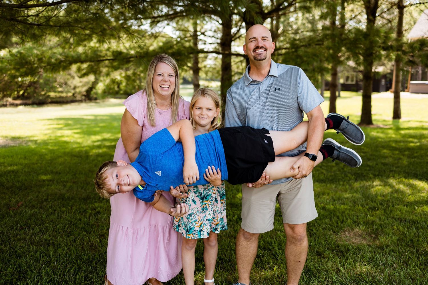 A cheerful family stands together on a sunny day in a grassy park with pine trees in the background.