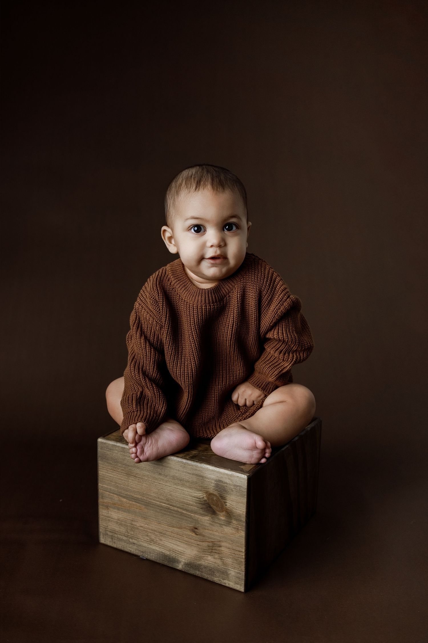 Baby in brown knit sweater sits on wooden box against dark background in minimalist portrait studio setting.