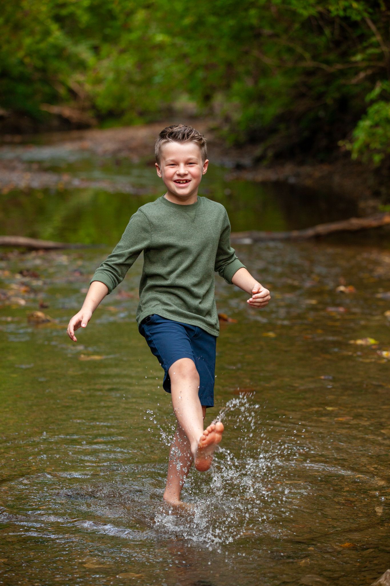 A joyful child in a green sweater and blue shorts splashes through shallow creek water on a sunny day.
