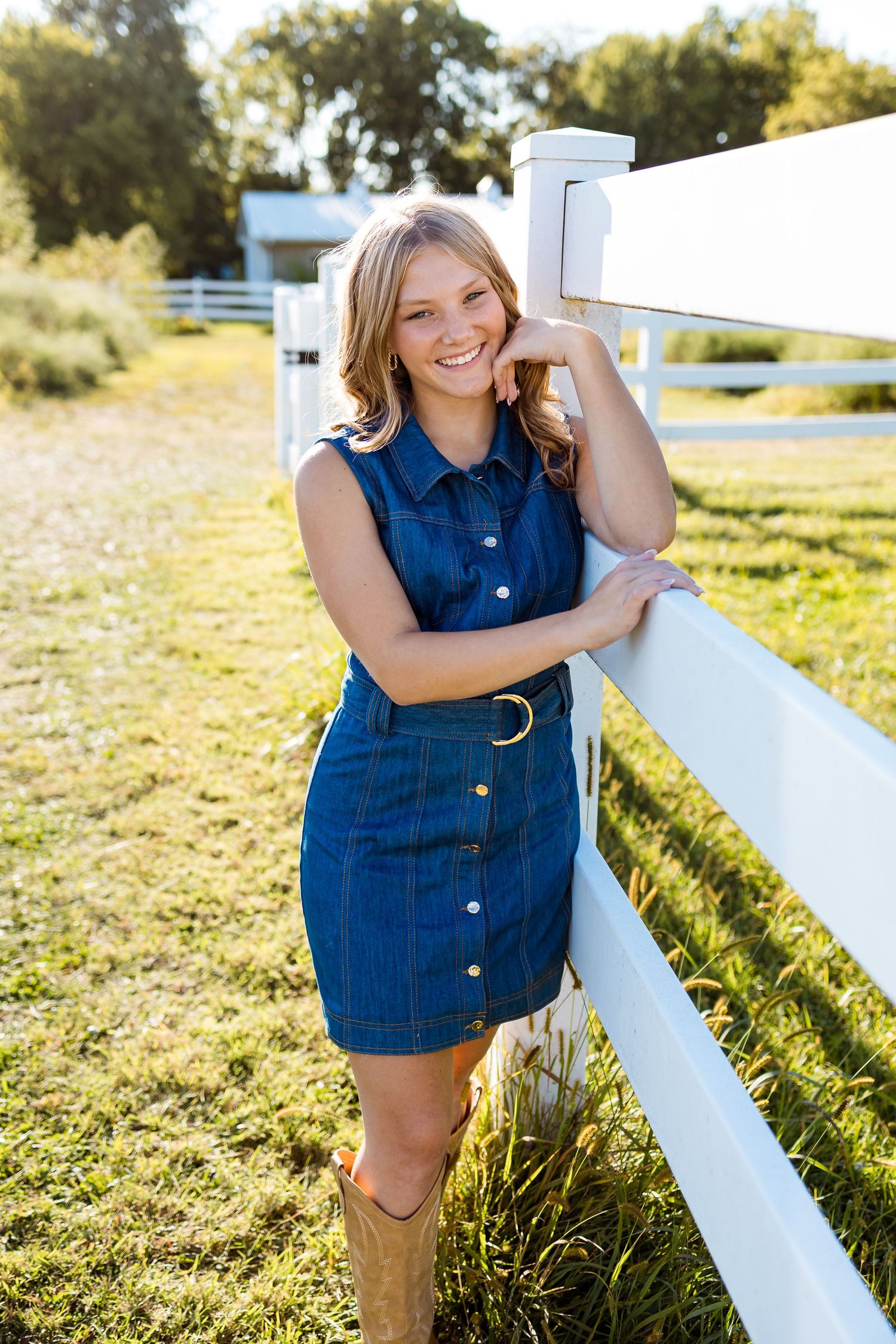 A denim button-down dress and cowboy boots create a casual country look against a white fence on a sunny day.