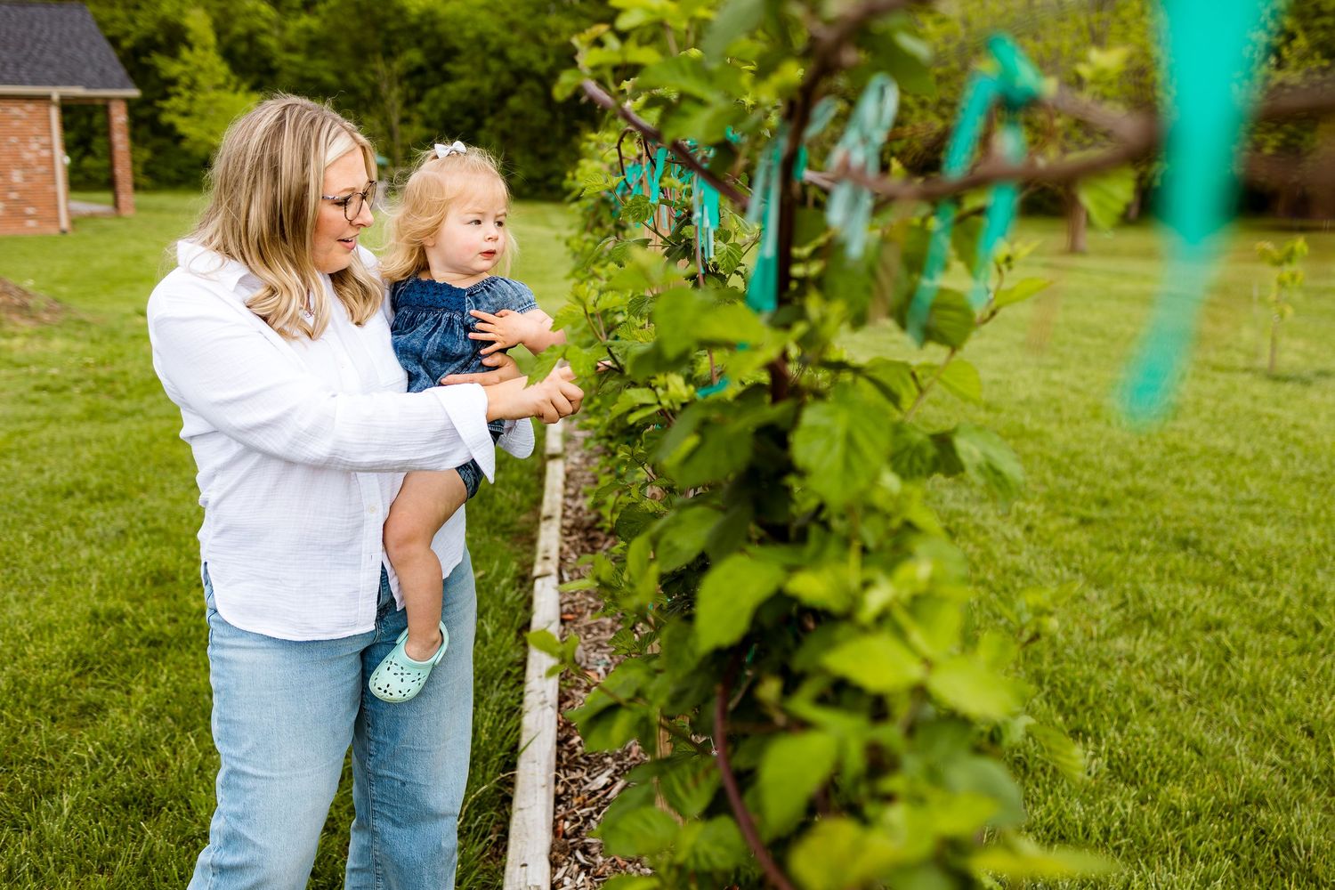 A parent helps a young child explore a garden in a backyard with green plants and trees.
