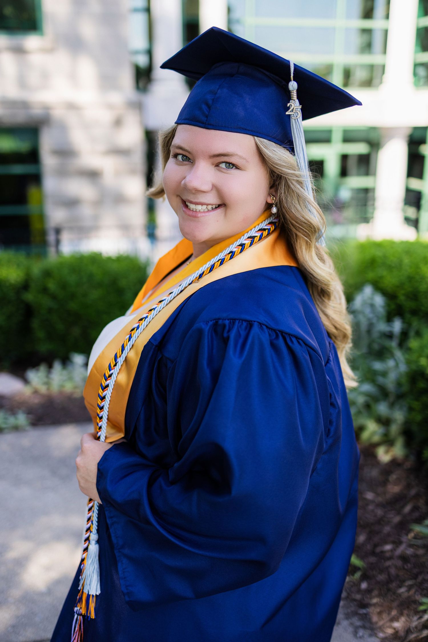 Graduate in blue cap and gown with gold stole and honors cords poses outdoors on graduation day.