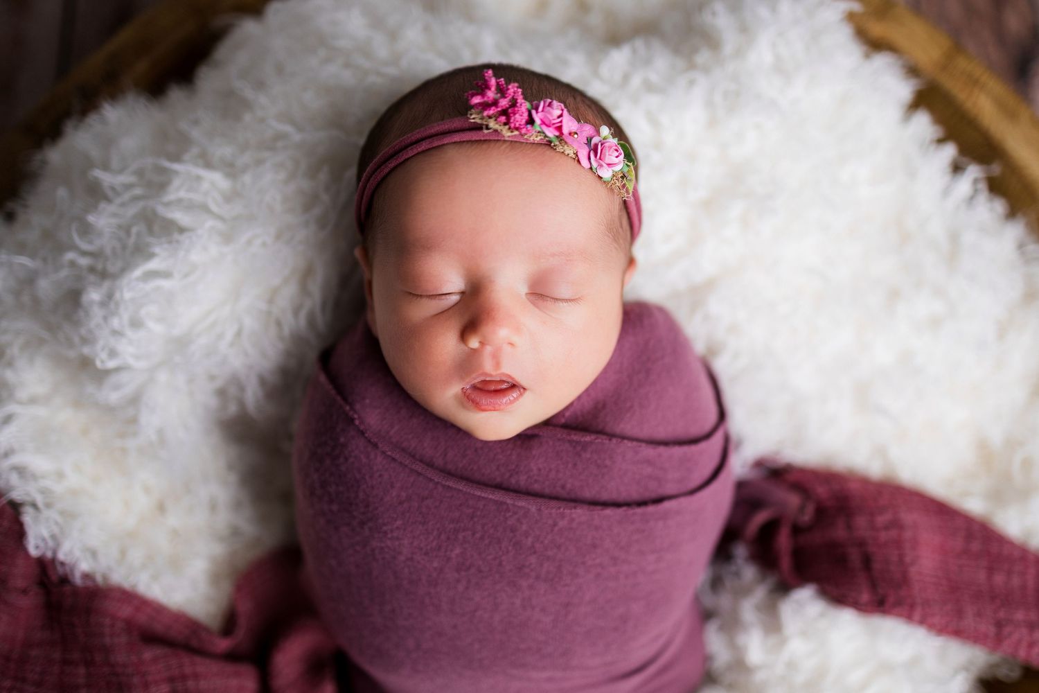 Newborn baby wearing pink flower headband sleeps peacefully wrapped in purple swaddle on white fluffy blanket.