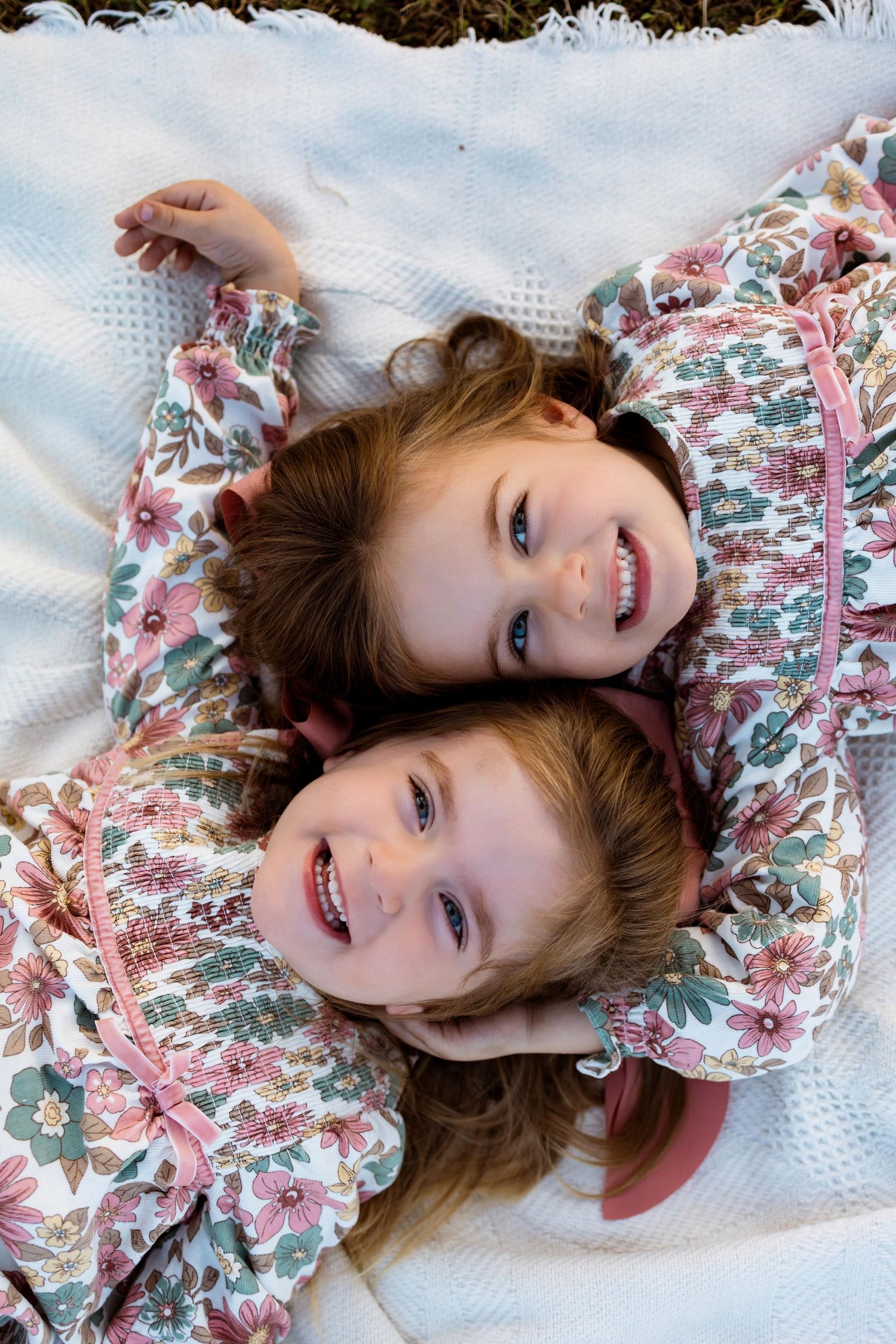 Children in floral dresses lay on white blanket during outdoor portrait session.