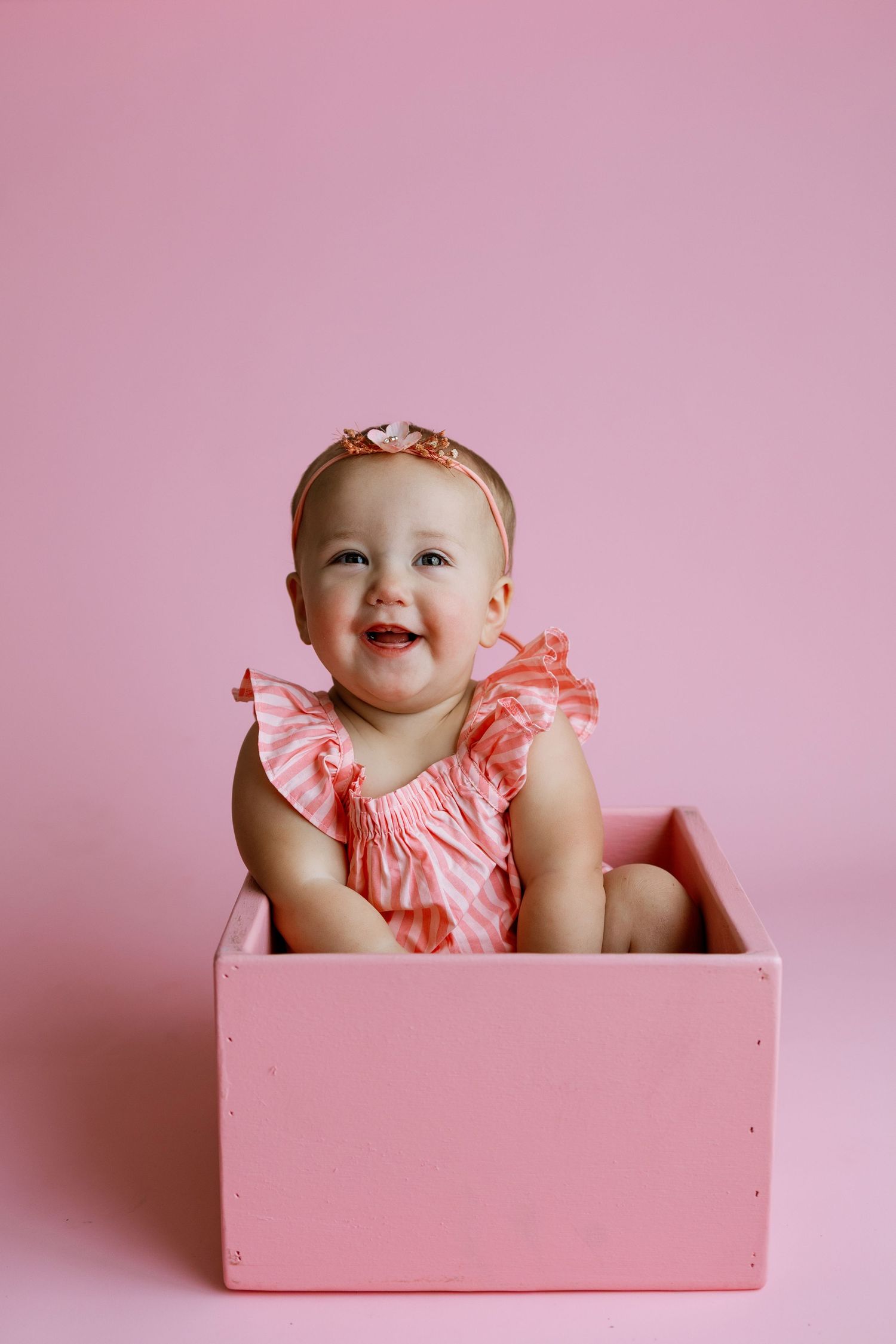 Smiling baby girl in pink ruffled romper and headband sitting in a pink wooden box against a pink background.