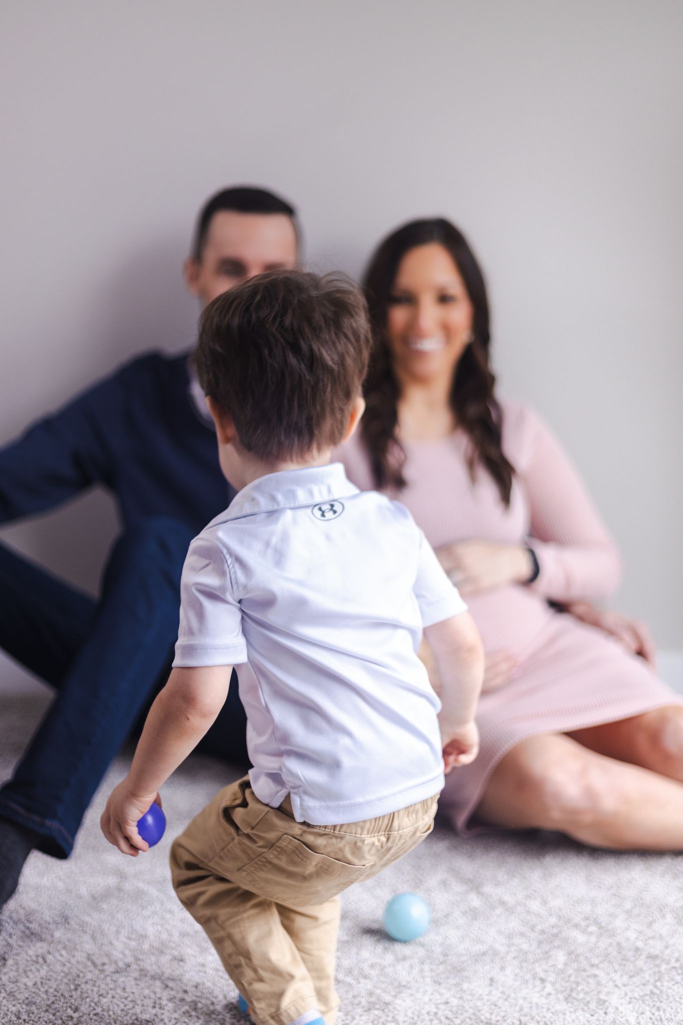 Child in white shirt and khakis playing with toys while parents watch from couch.