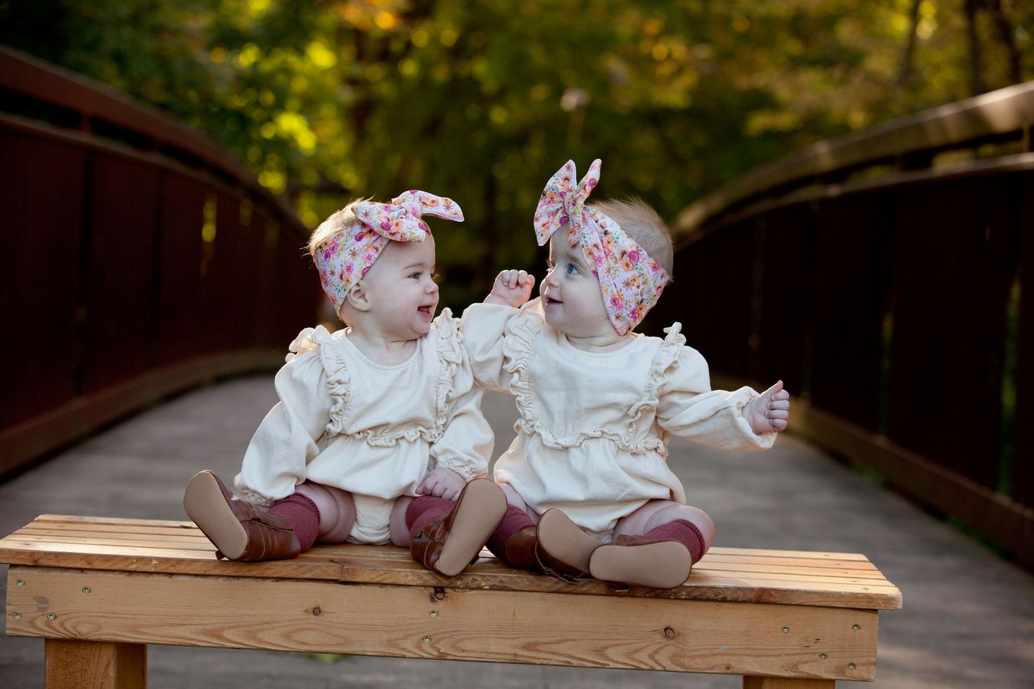Two babies in matching white dresses and purple bows sit together on a wooden bench in a park setting.