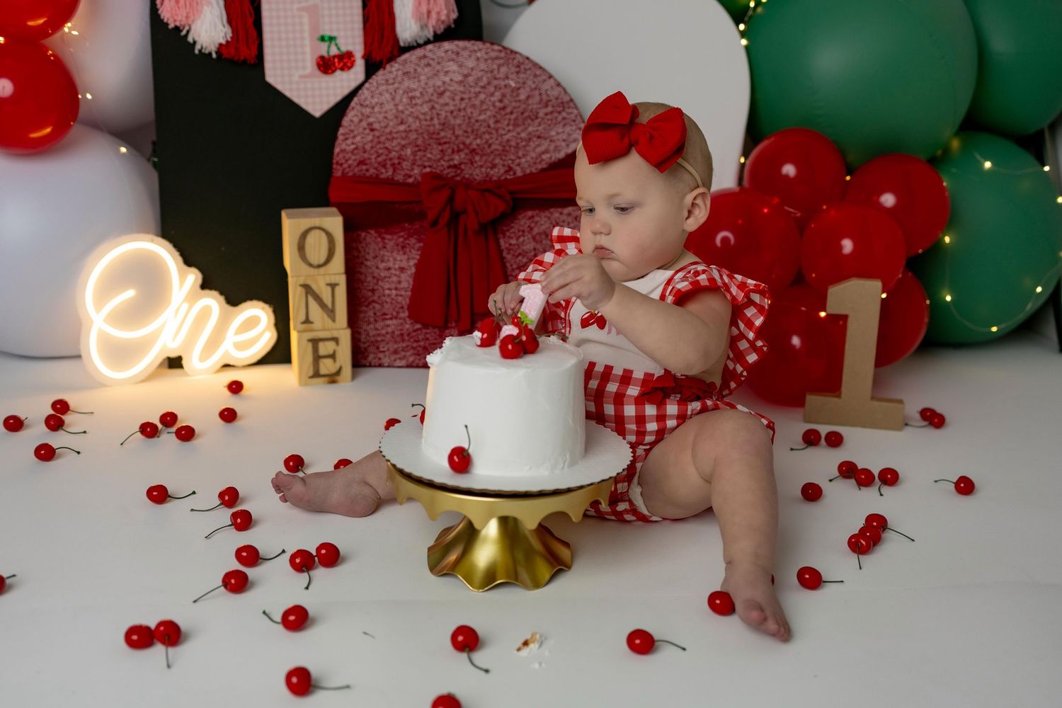 Cherry-themed birthday setup with red and green balloons, blocks, and gingham outfit details.