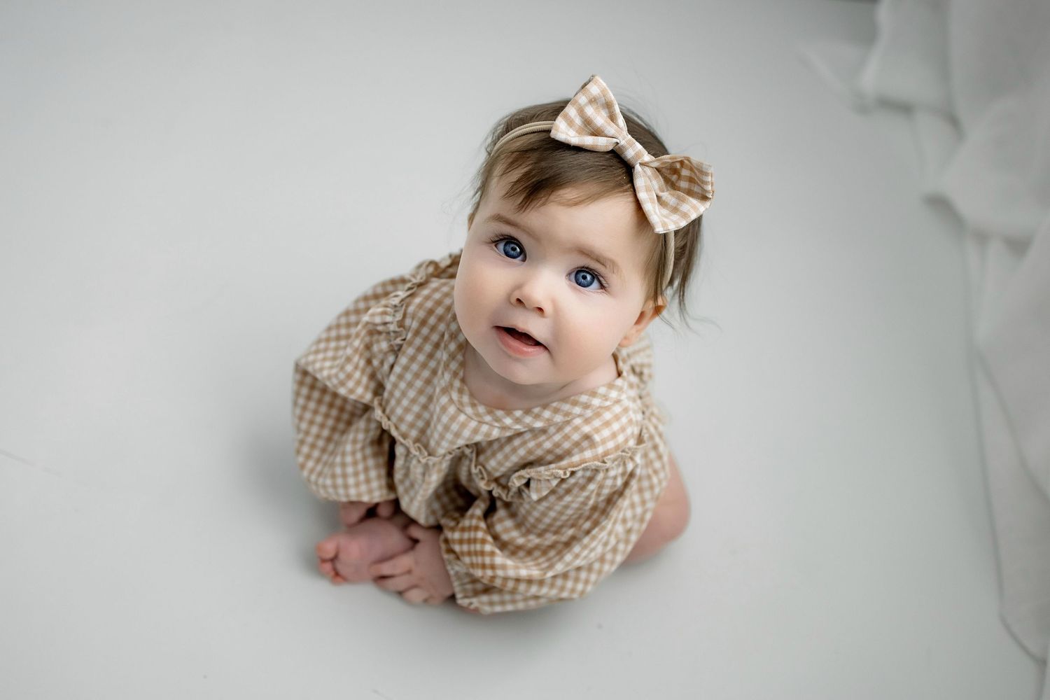 Baby wearing a beige knit sweater and bow headband lies on soft white bedding while looking upward.