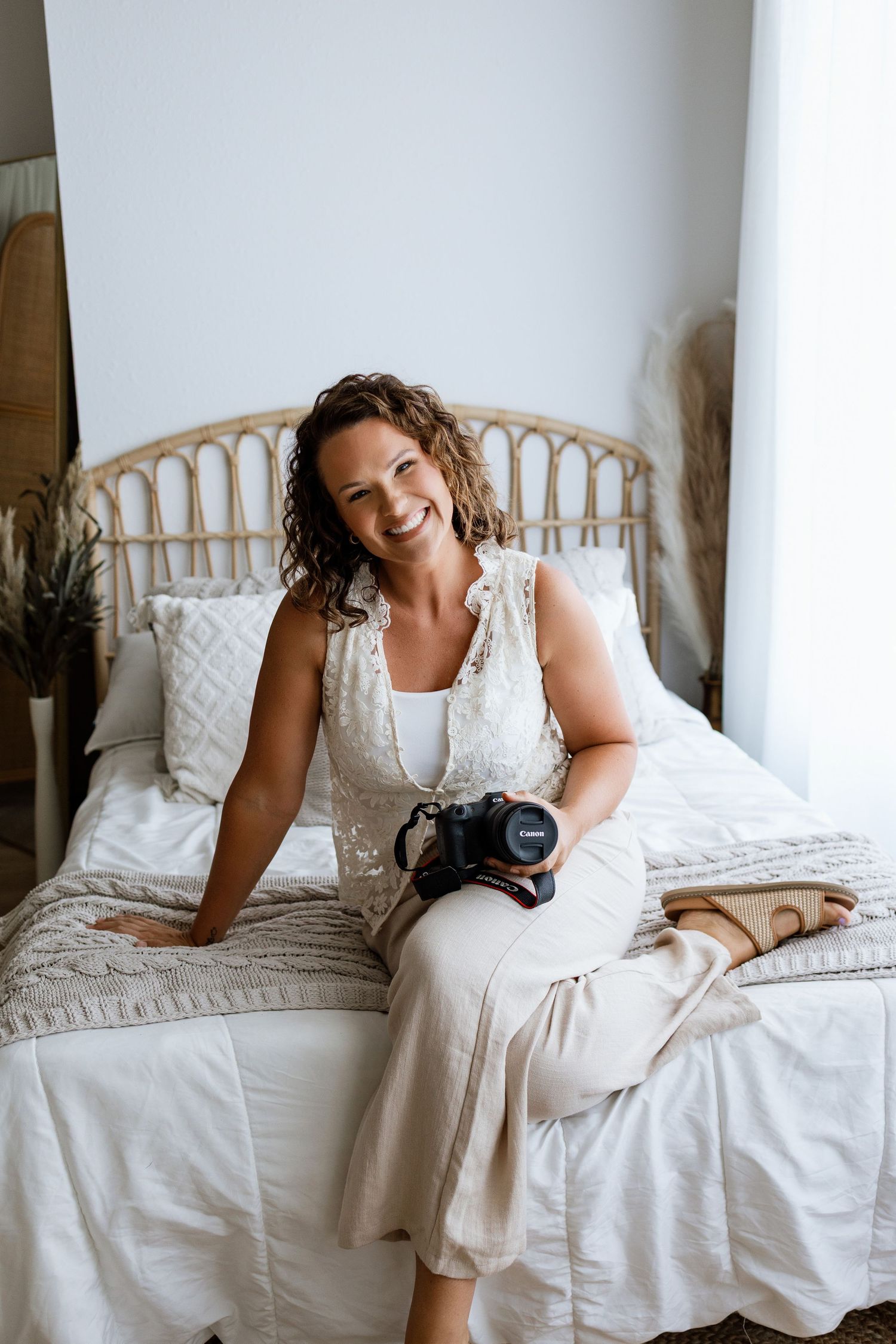 Smiling woman in white dress sitting on bed holding Canon camera in bright boho bedroom setting.