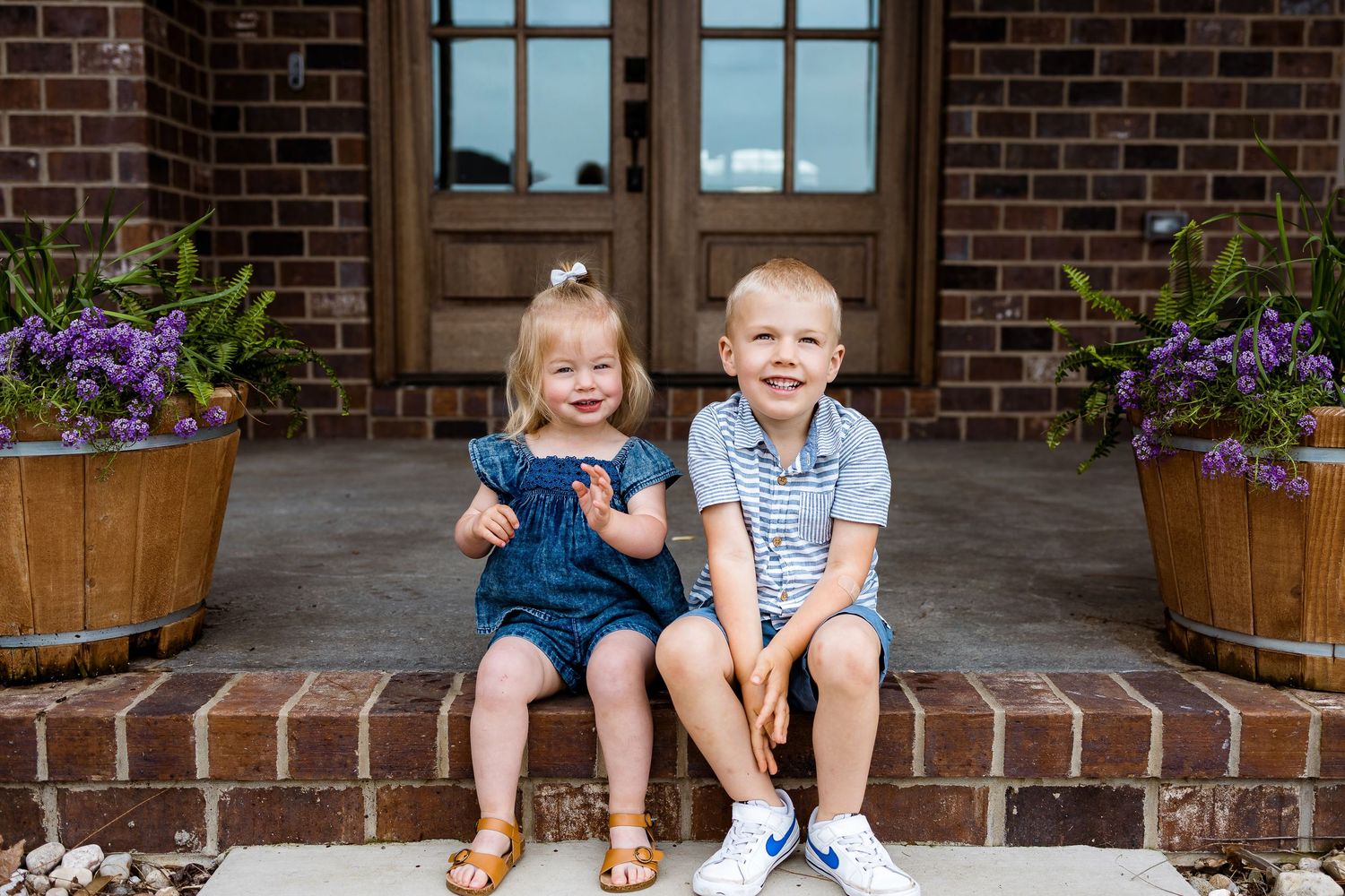 Two children sitting on brick steps between planters with purple flowers share a playful moment.
