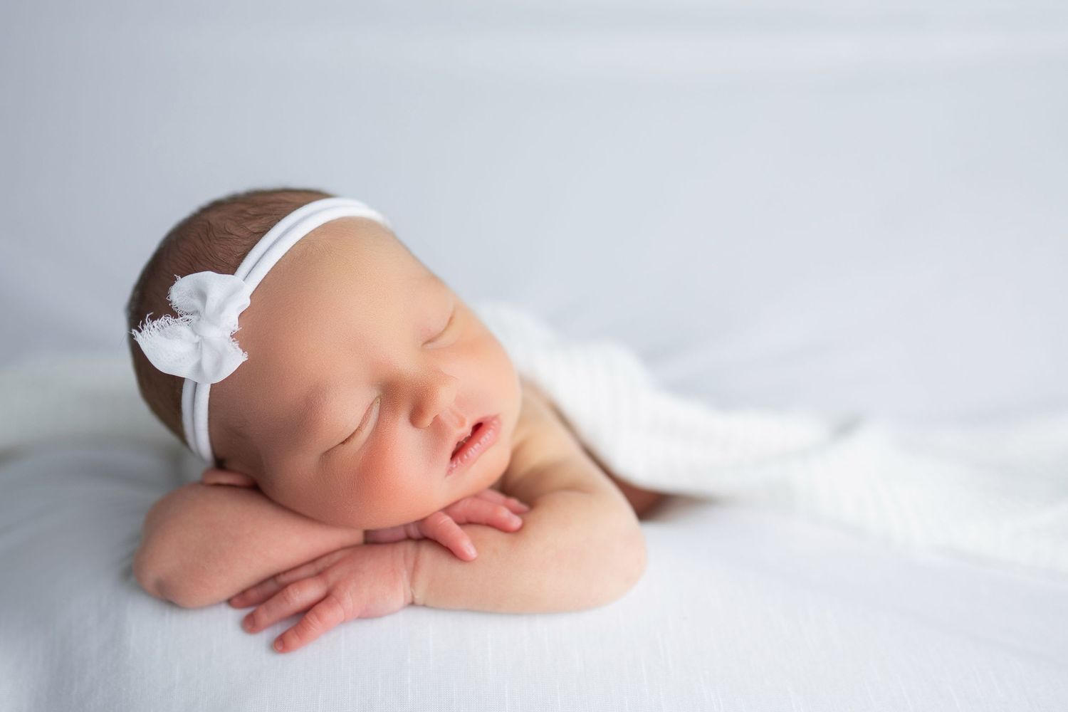 A sleeping baby wrapped in soft white fabric wearing a delicate white bow headband laying on their hands in a gentle pose.
