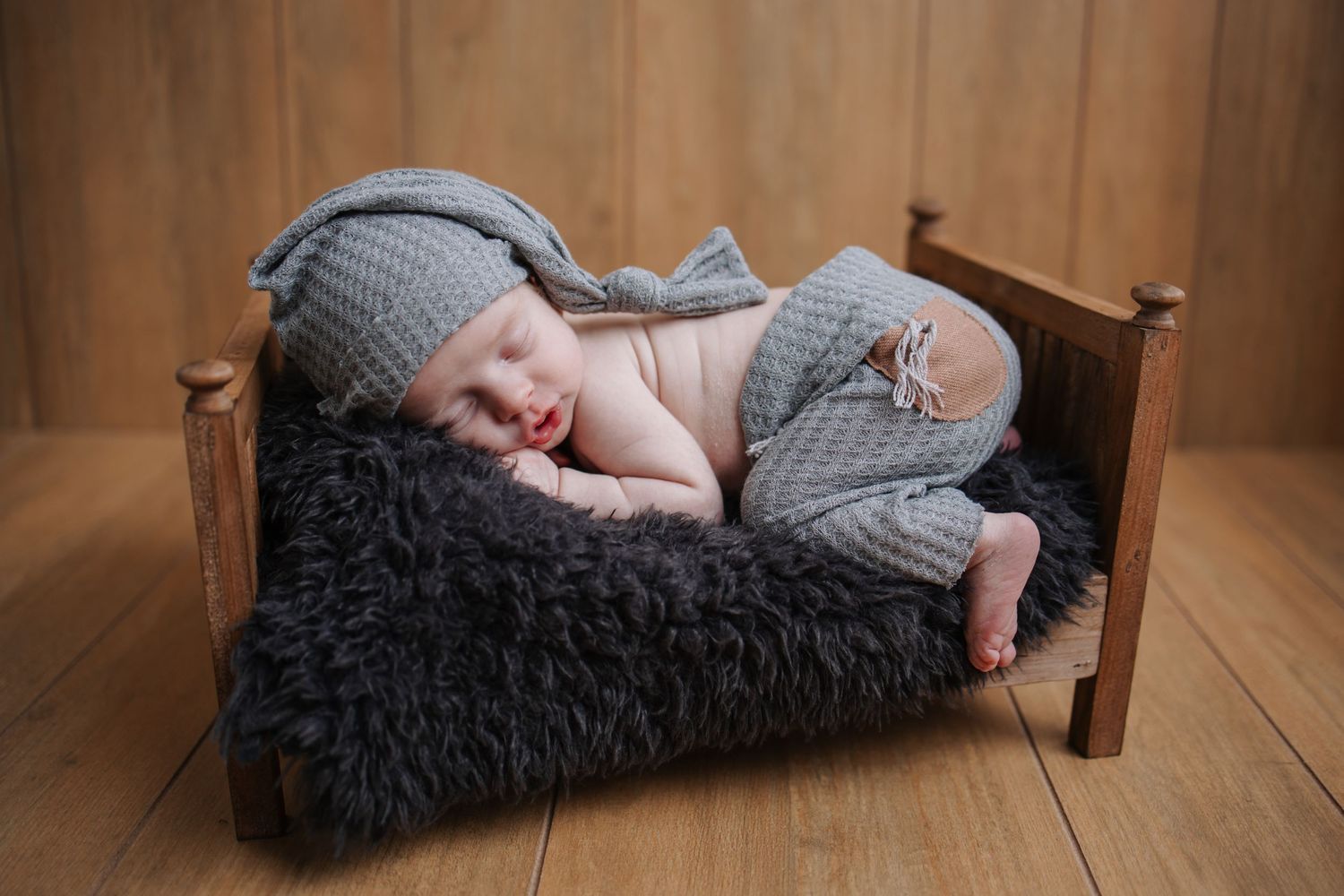 Sleeping newborn on miniature wooden bed with grey knit outfit.