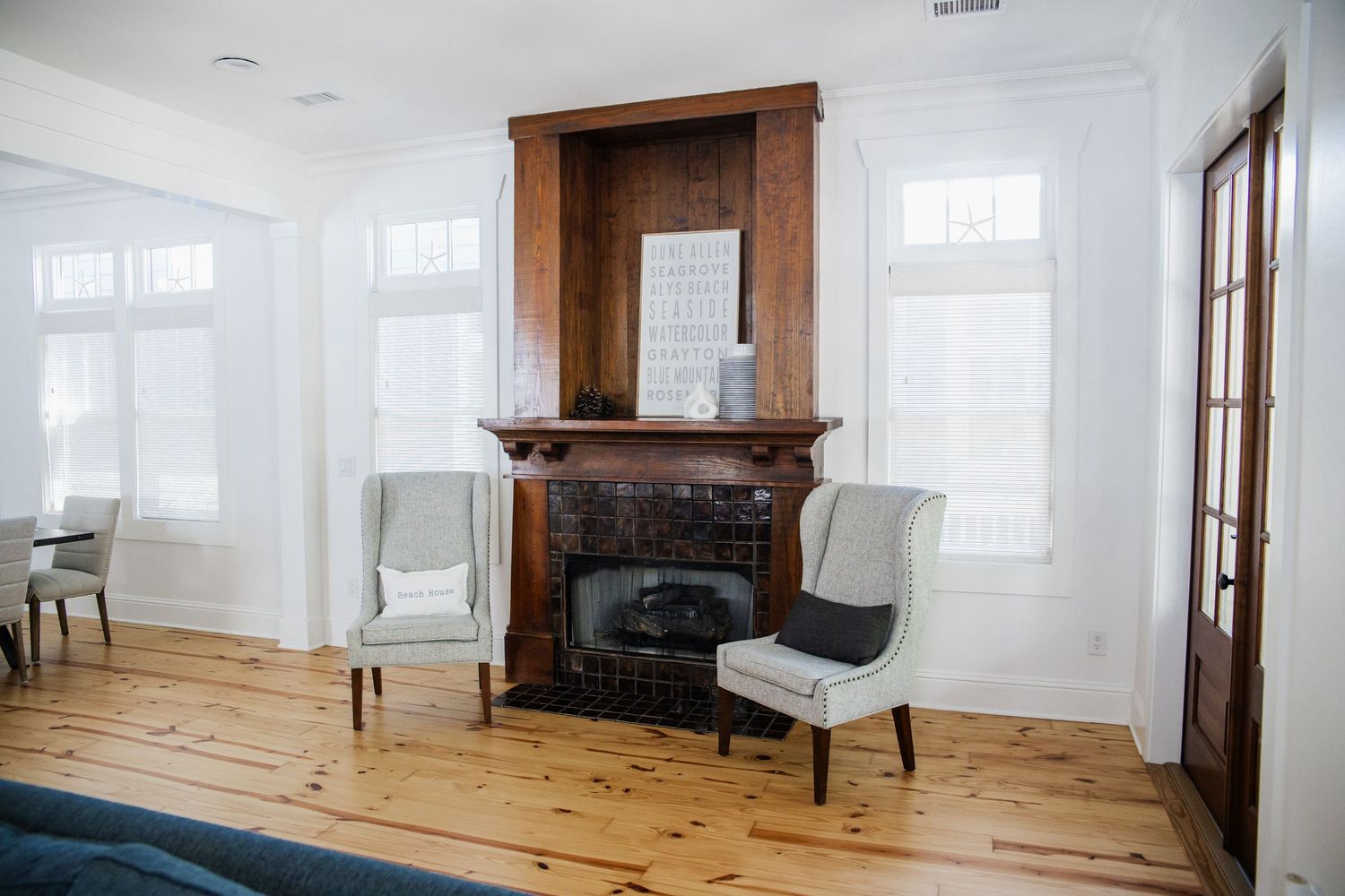Classic wooden fireplace mantel with white wingback chairs in bright room with pine flooring.