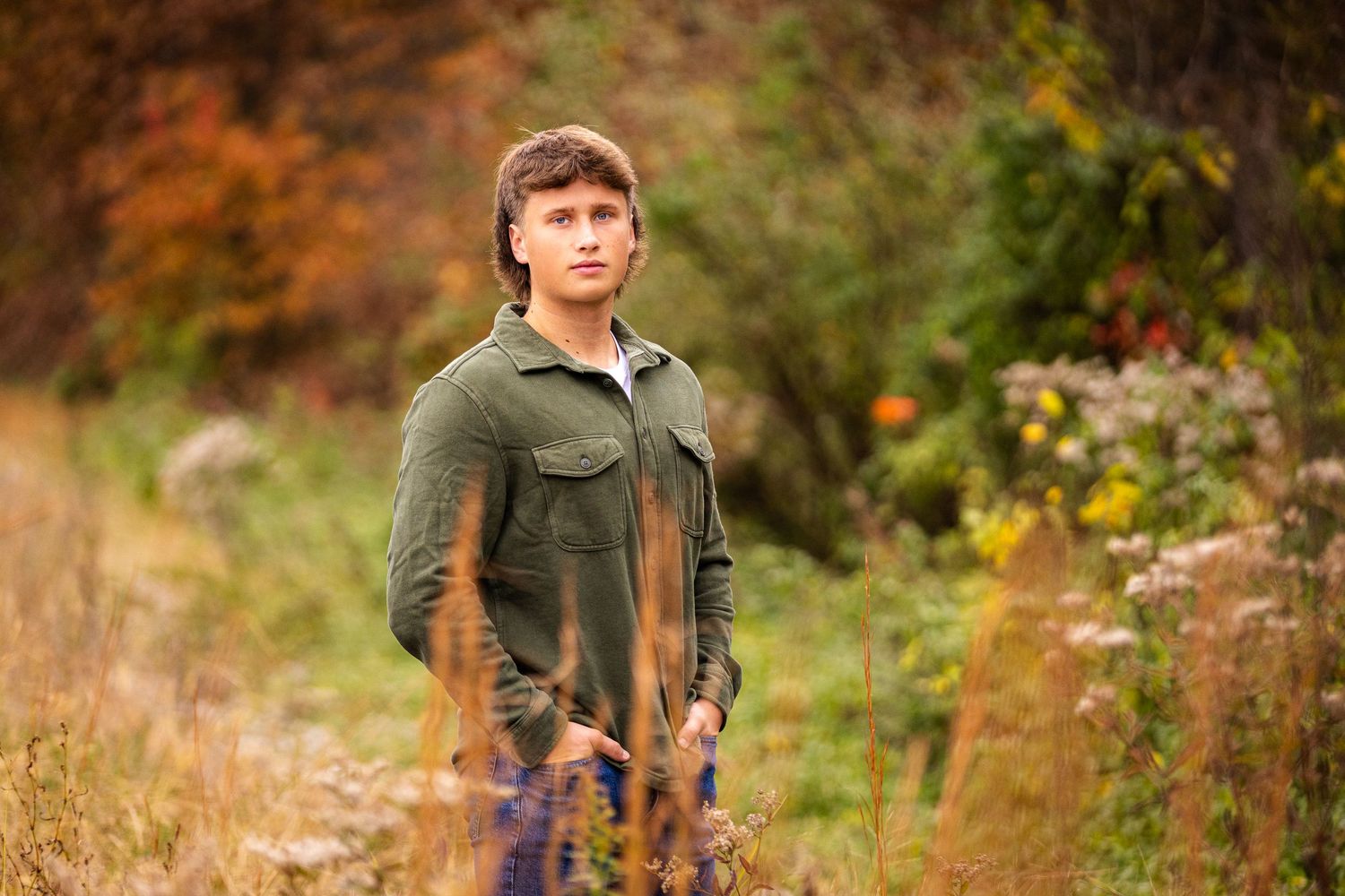 Person in green jacket standing in field with tall grass and fall colors.