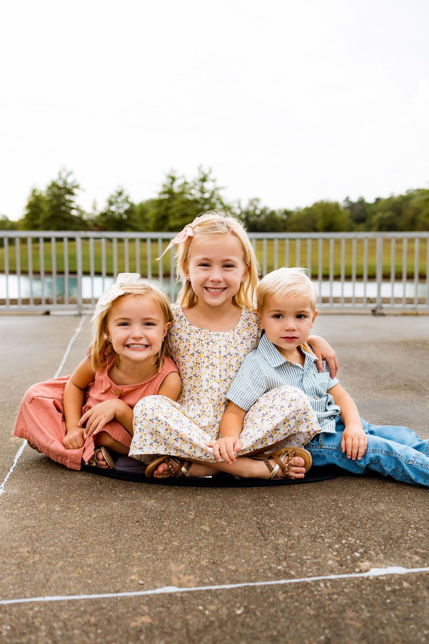 Three children lie on the ground together in a row wearing floral dresses and casual clothing outdoors.