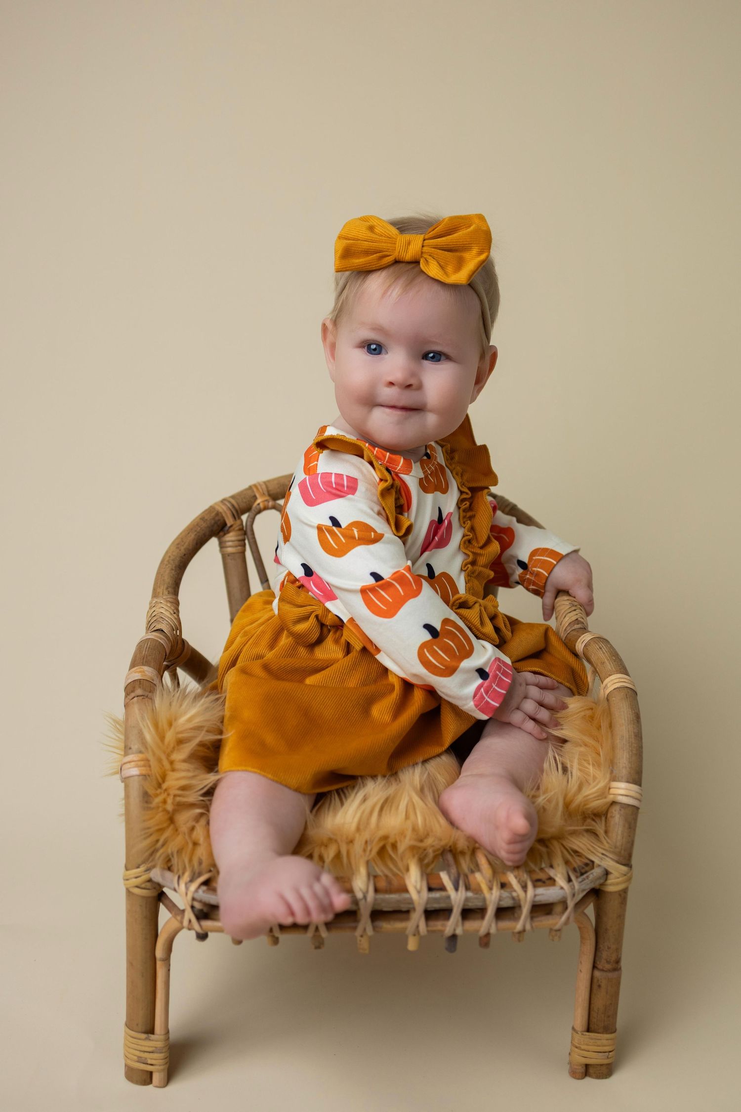 Baby in orange dress with pumpkin print sits on rustic bamboo bench in fall-themed photography session.
