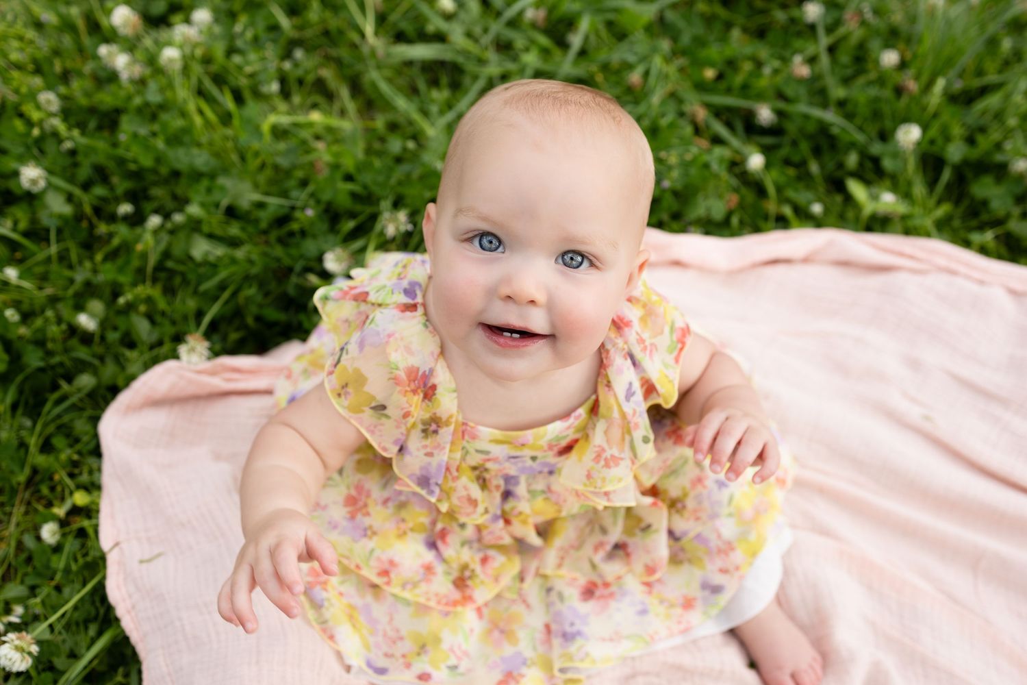 Happy baby in floral dress sitting on pink blanket surrounded by green grass and flowers.
