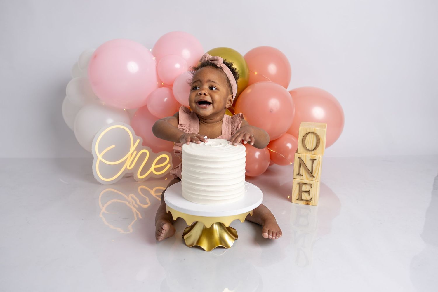 Smiling baby reaches for white birthday cake surrounded by pink and gold decorations.