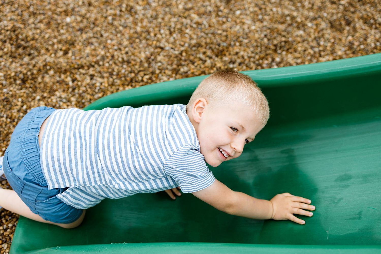 Child playing and sliding down a green playground slide over wood chips on a sunny day.