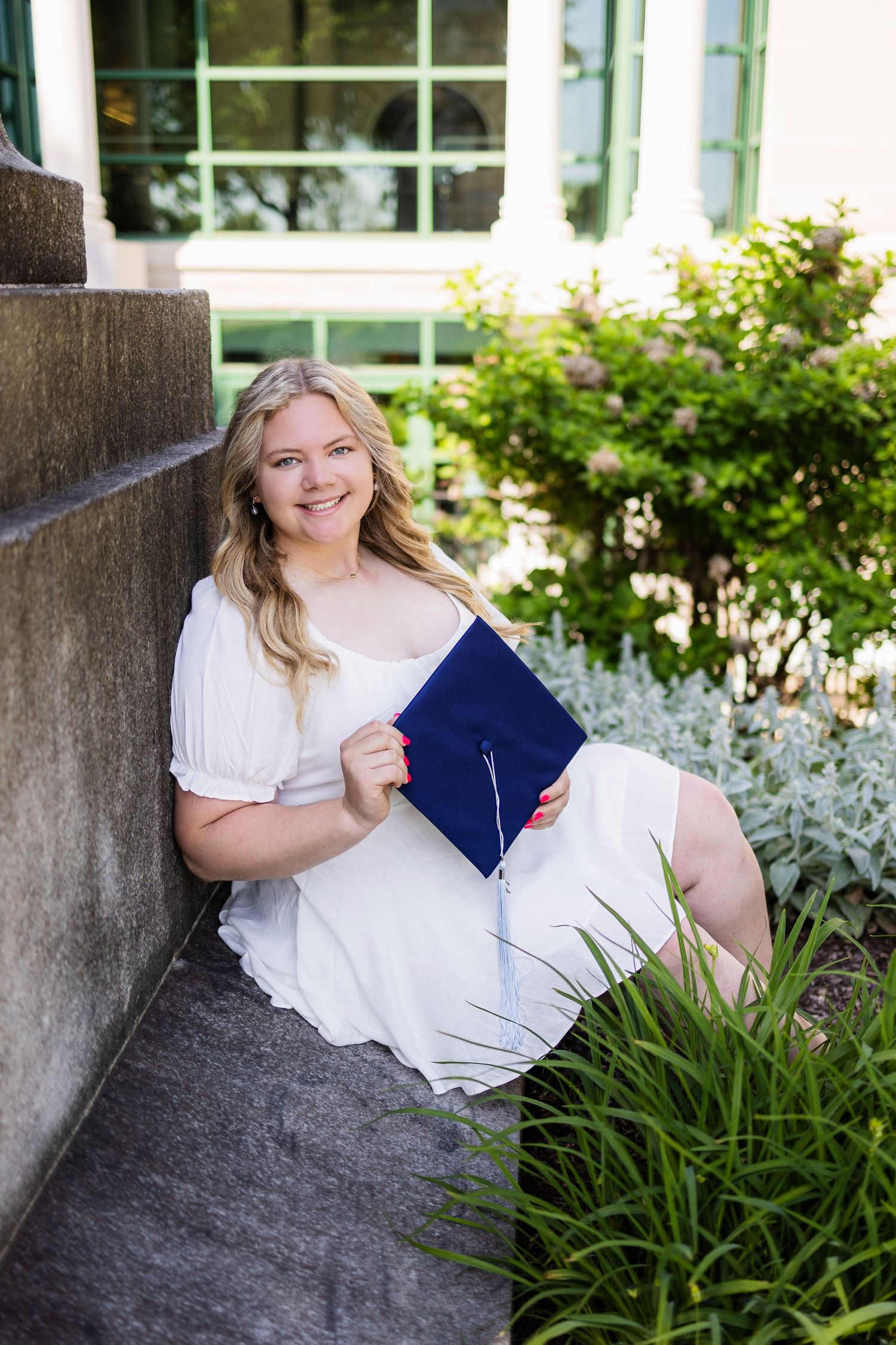 A graduate in a white dress poses with diploma holder while sitting on stone steps among landscaping.