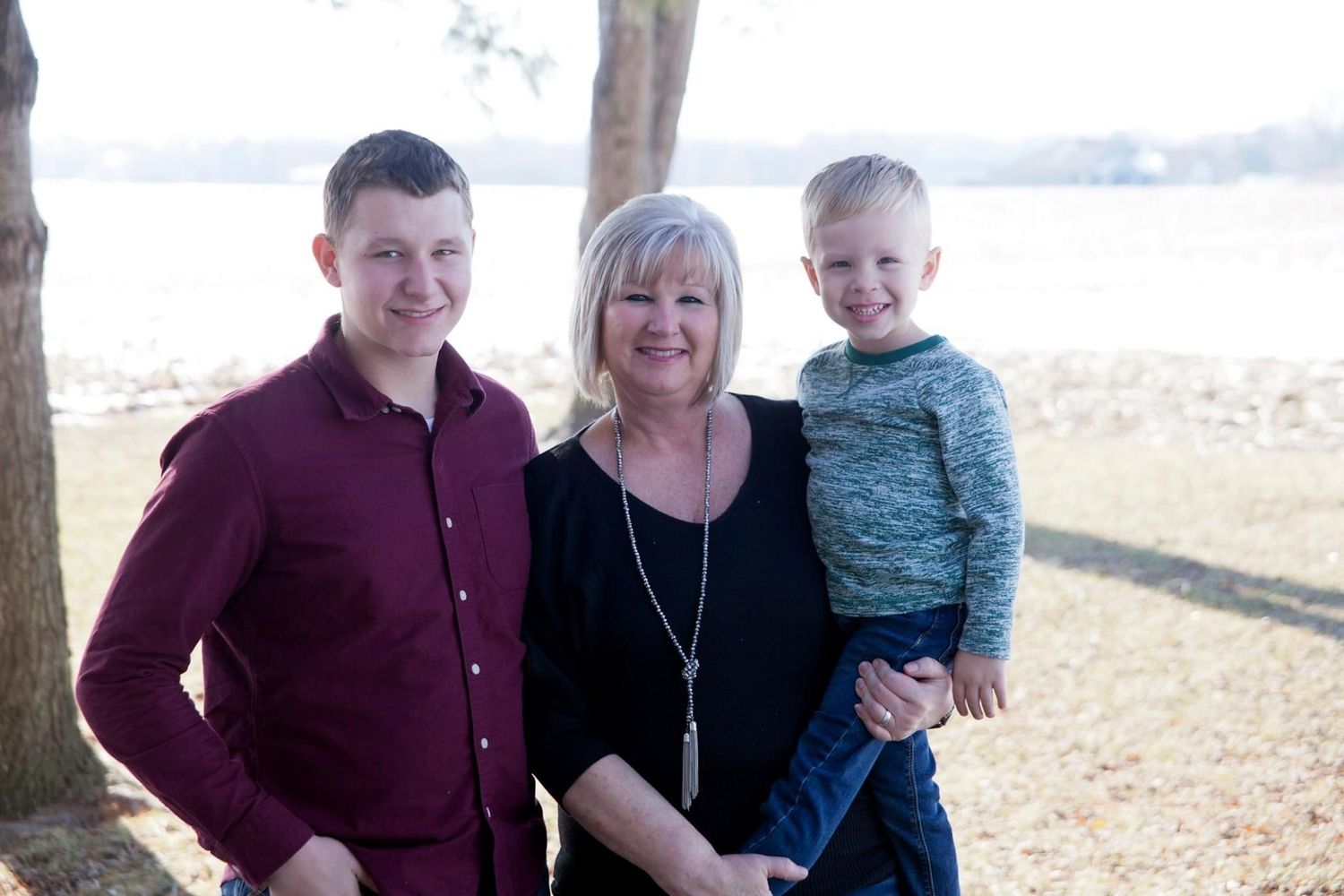 A woman smiles outdoors near a lake, posing with a teenage boy and a young child in a warm family portrait.