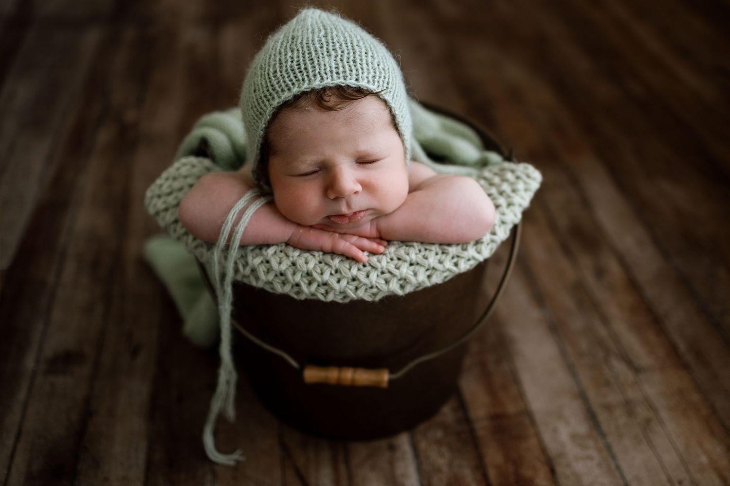 Sleeping newborn baby in green knit bonnet and wrap posed in a wooden bucket on dark hardwood floors.