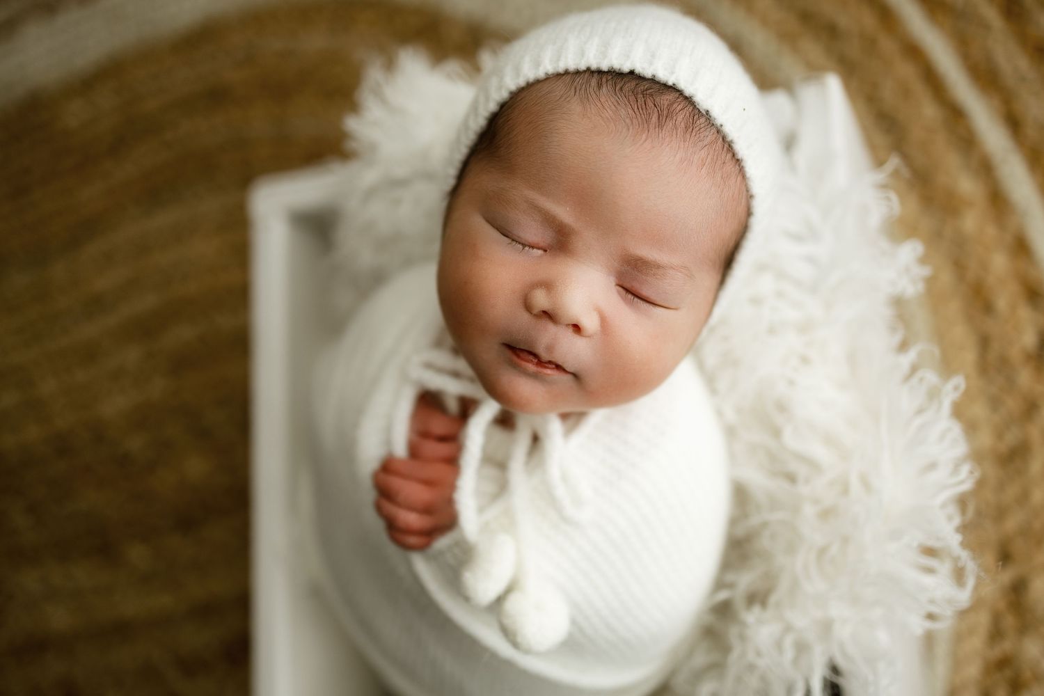 Sleeping newborn baby wrapped in white blanket with feathery texture against warm brown background.