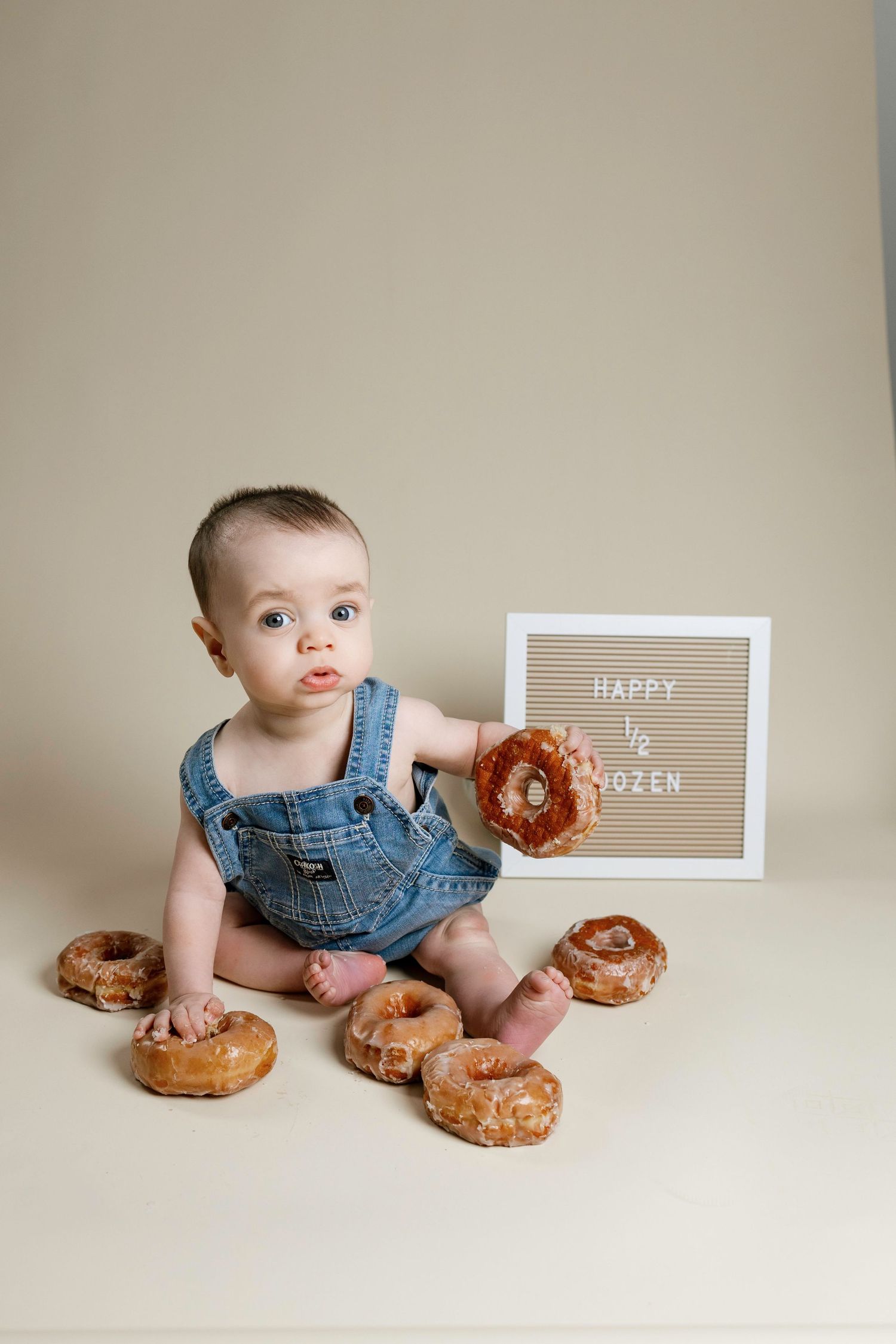 Baby in denim overalls sits on beige backdrop with donuts and wooden signs in a series of playful poses.