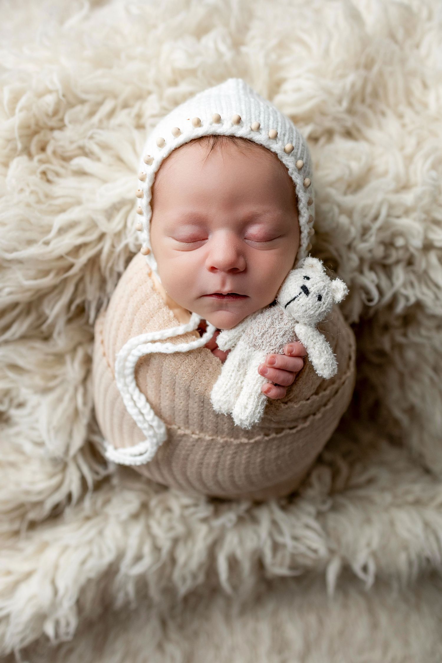 Sleeping newborn wrapped in cozy beige knit blanket on fluffy white fur background in professional portrait series.