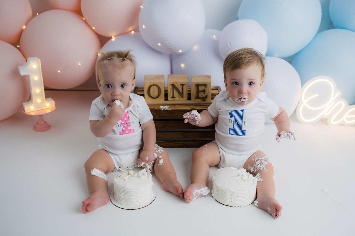 First birthday celebration with twins sitting on white cushions surrounded by pink and blue balloons.