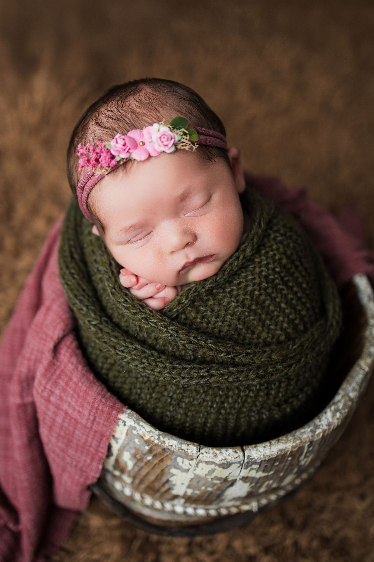 Sleeping newborn wrapped in olive green knit blanket with pink flower headband in rustic container.